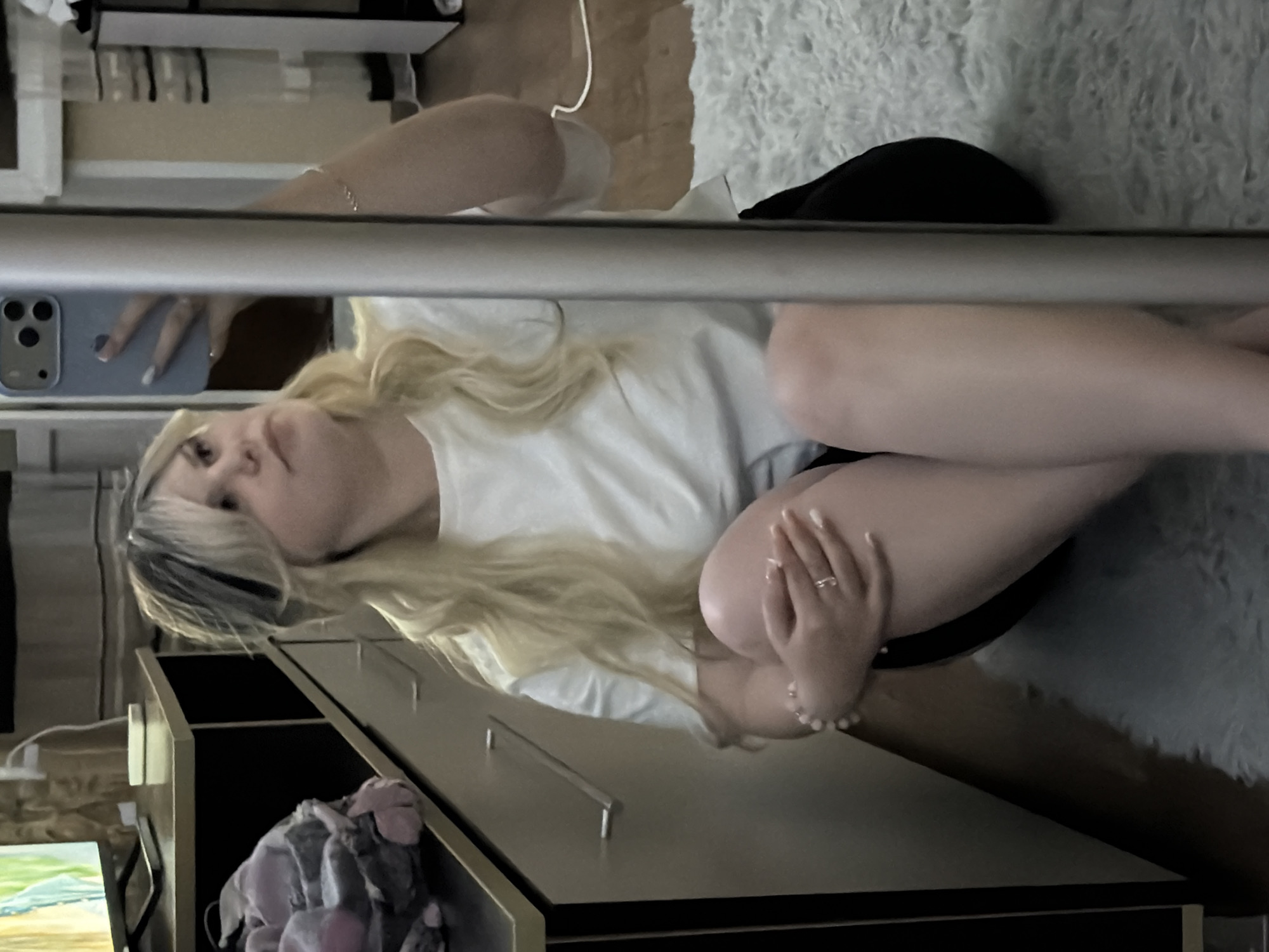 A young woman is sitting on a light-colored rug in what appears to be a bedroom. She is holding a light blue smartphone and taking a selfie in a mirror. She has long blonde hair with some darker streaks and is wearing a white t-shirt and black shorts. A piece of furniture with drawers is visible to her left, with a television on top. The background shows a wooden floor and a white cord from an electrical appliance. The woman is wearing a ring and a bracelet. The overall impression is a casual, personal moment captured in a home environment. The location is specified as Belgorod, Russia, though there are no specific visual cues within the image to confirm this.