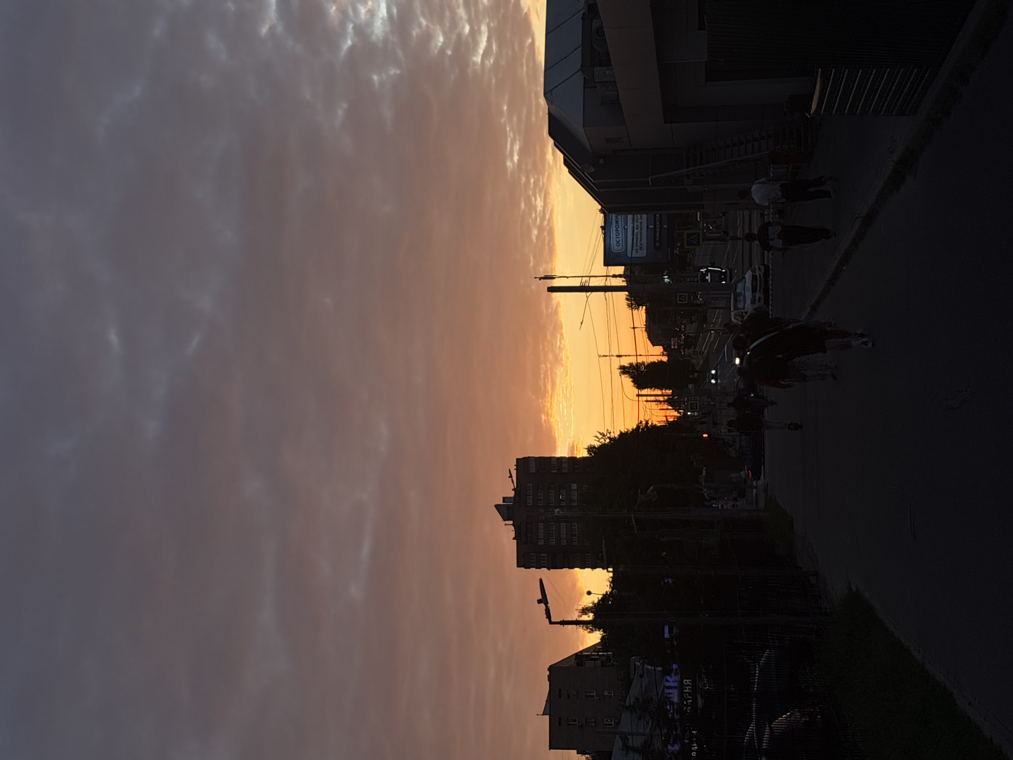 This media file depicts an urban street scene during sunset. The sky is filled with a gradient of warm colors, transitioning from pale orange near the horizon to a muted purple and gray higher up, with scattered clouds. Silhouettes of buildings and trees are visible against the colorful sky.
In the foreground and middle ground, several people are walking along a sidewalk and a street. One person is riding a bicycle. Cars are also present on the street. The setting appears to be a city, with apartment buildings and what looks like a commercial structure with a large ribbed facade. Streetlights and power lines are also visible.
The time of day is clearly sunset, characterized by the dramatic sky and the long shadows. The weather appears to be clear, with the clouds contributing to the visual spectacle of the sunset. The overall mood conveyed by the image is calm and transitional, reflecting the end of the day.
Visible text includes "ОСТОРОЖНО" on a blue and white sign, which translates to "CAUTION" in Russian. Another sign reads "YUGACENTER". There are also other signs with Cyrillic text, but they are not clearly legible.