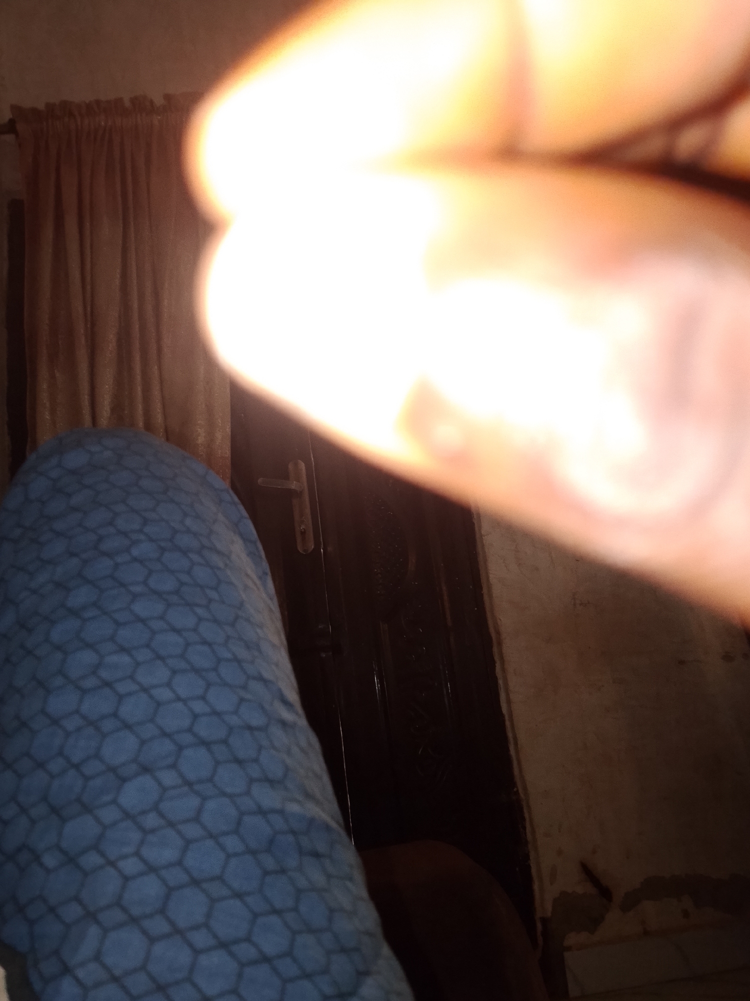 The image shows a close-up view of a person's leg covered by a blue fabric with a hexagonal pattern. The fabric is wrinkled and appears to be a blanket or a soft garment. To the right of the leg, a dark door with a silver handle is visible. Behind the leg and to the left, brown curtains are drawn, suggesting an indoor setting. A bright light source, possibly a flash or a strong light from a window, is creating an overexposed area in the upper right portion of the image, blurring out any details in that region. The lighting suggests it might be daytime, though the indoor setting makes it difficult to determine definitively. The overall impression is of a casual, domestic scene.