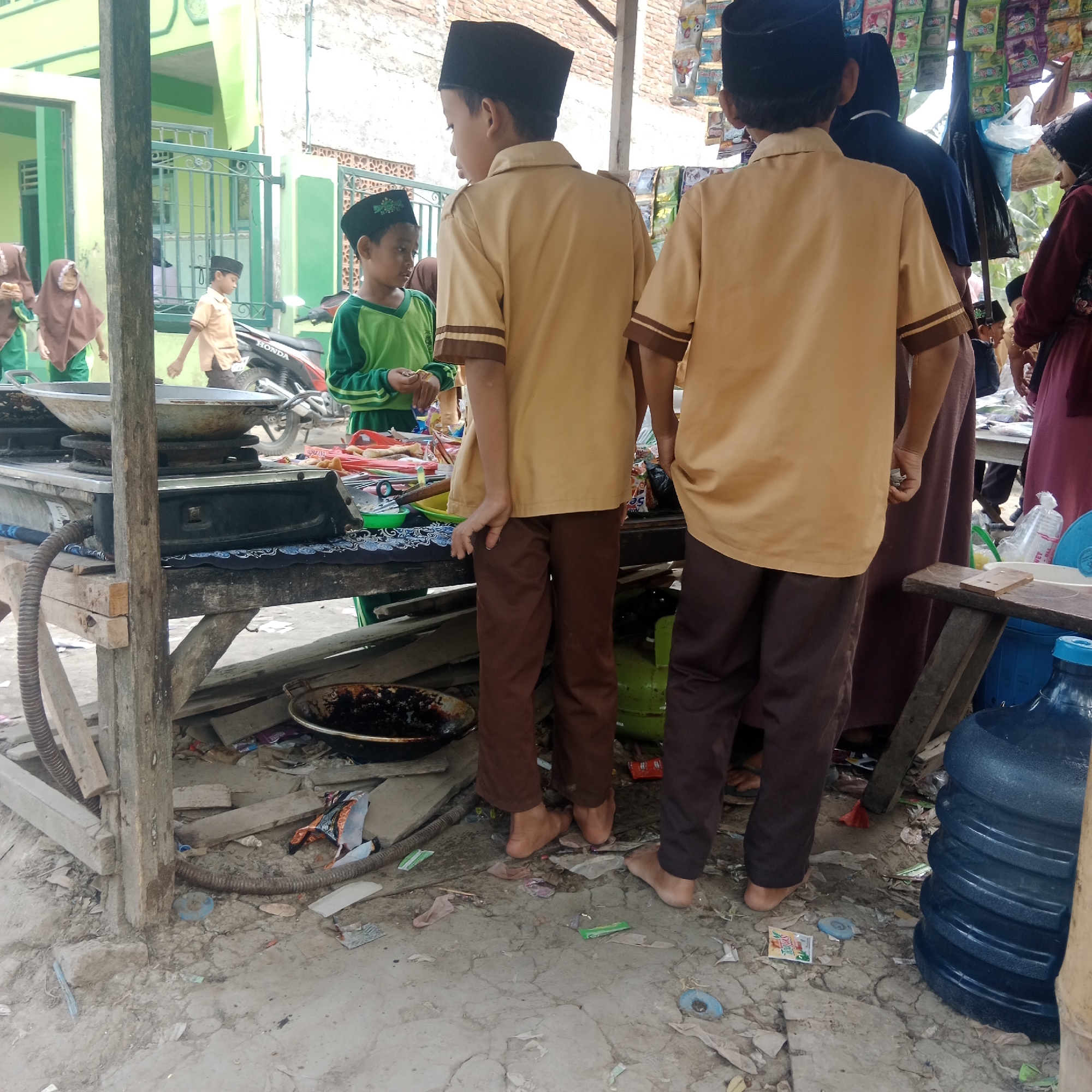 In Tegowanu Kulon, Indonesia, a scene unfolds at an outdoor food stall with two young boys wearing light brown school uniforms and black peci caps standing. The boy on the left wears a green long-sleeved shirt under his uniform, while the boy on the right is in full brown uniform. They are positioned near a makeshift stall constructed from wood and covered with a tarp. A gas stove with a large wok is visible on the stall, suggesting cooking activities. In the background, a motorcycle is parked, and other individuals, including two young girls in headscarves and a woman in a dark hijab and maroon dress, are present. The ground is littered with debris, including plastic wrappers, broken pieces of wood, and a large blue water jug. The stall itself displays various packaged snacks and food items. The overall environment appears to be a busy, informal street setting.