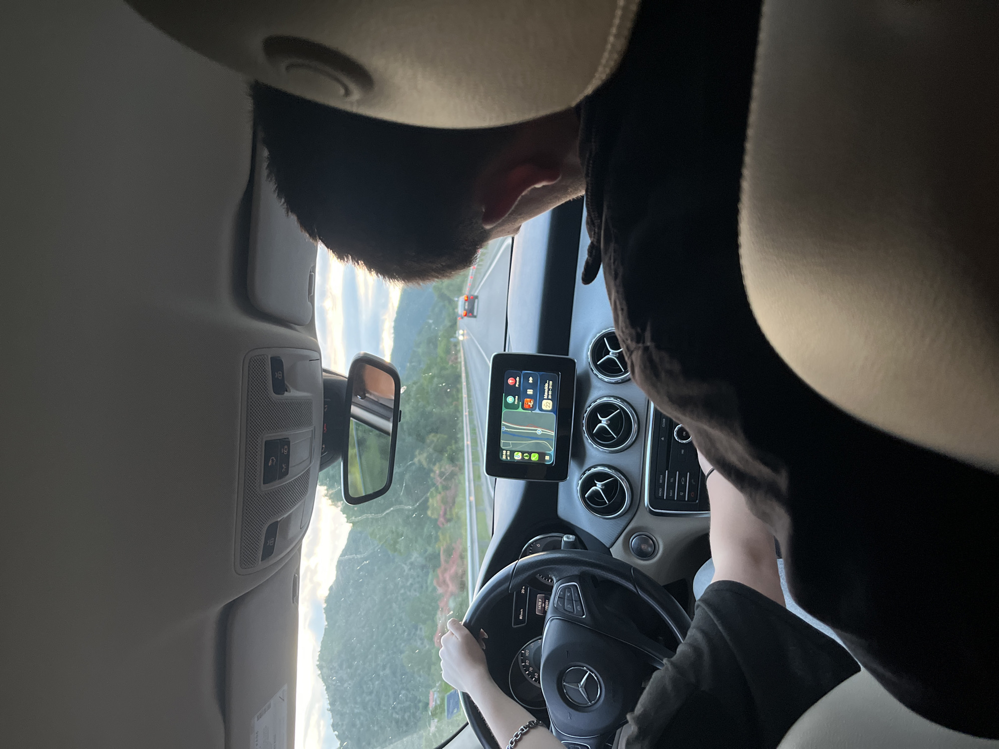 The image shows the interior of a car driving on a road in an area with green hills. The dashboard, steering wheel, and center console of the car are visible. The driver is holding the steering wheel and there is a navigation screen showing a map. The road ahead is visible through the windshield, with another vehicle in the distance. The location is Unknown, Unknown.