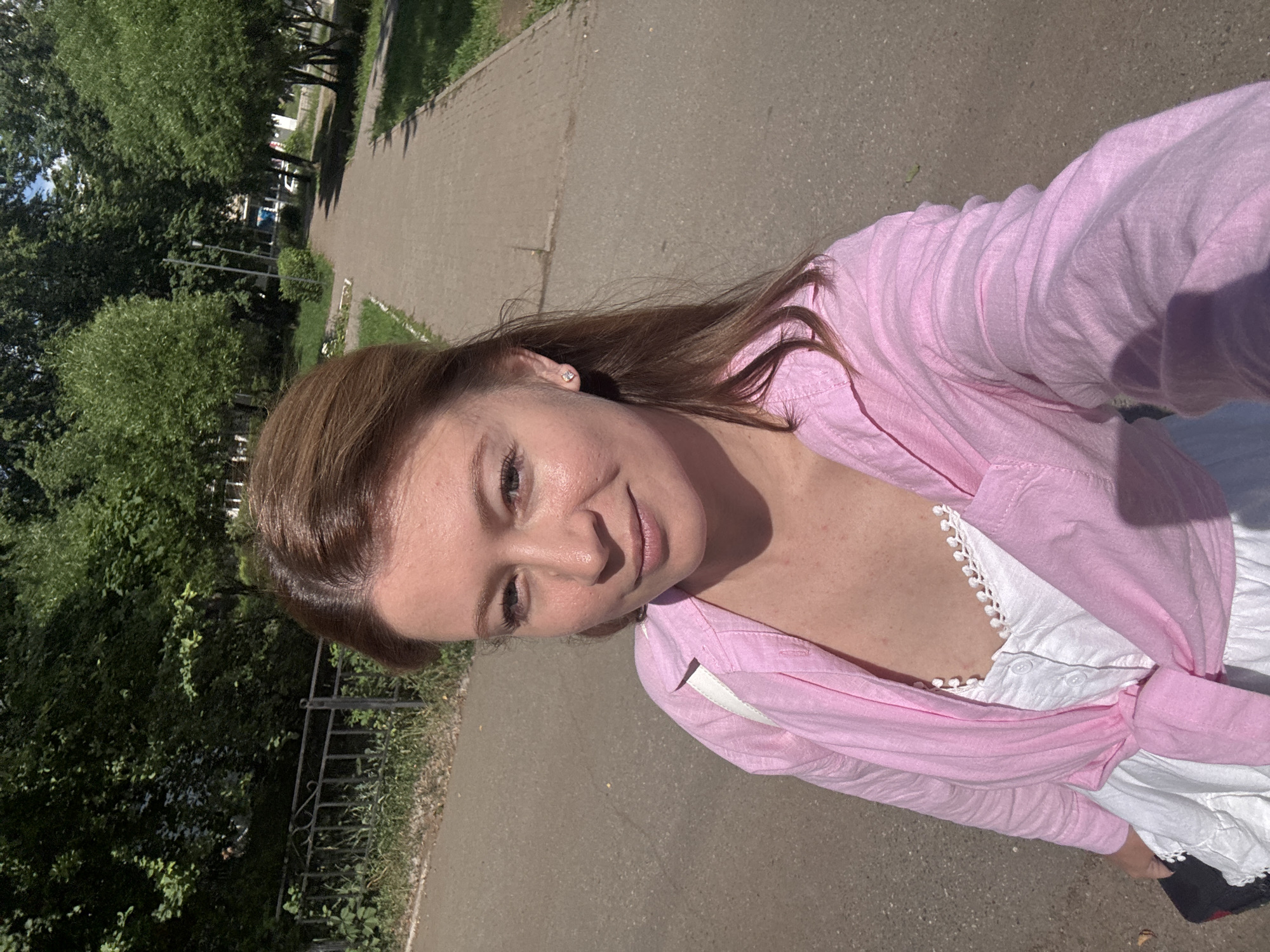 A young woman with shoulder-length brown hair takes a selfie outdoors on a sunny day. She is wearing a white camisole with decorative trim and a pink button-up shirt, which is tied around her waist. She has on small stud earrings. The background shows a paved walkway lined with green grass and trees. A metal fence is visible to the left. The scene appears to be a park or residential area in the city of Chaikovsky, Russia.