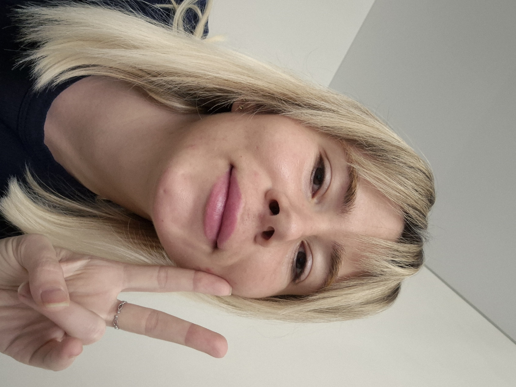 A woman with blonde hair is pictured close-up, tilting her head downwards and giving a peace sign with her left hand. She is wearing a dark-colored top. There is a white wall in the background. No specific location is discernible in the image.