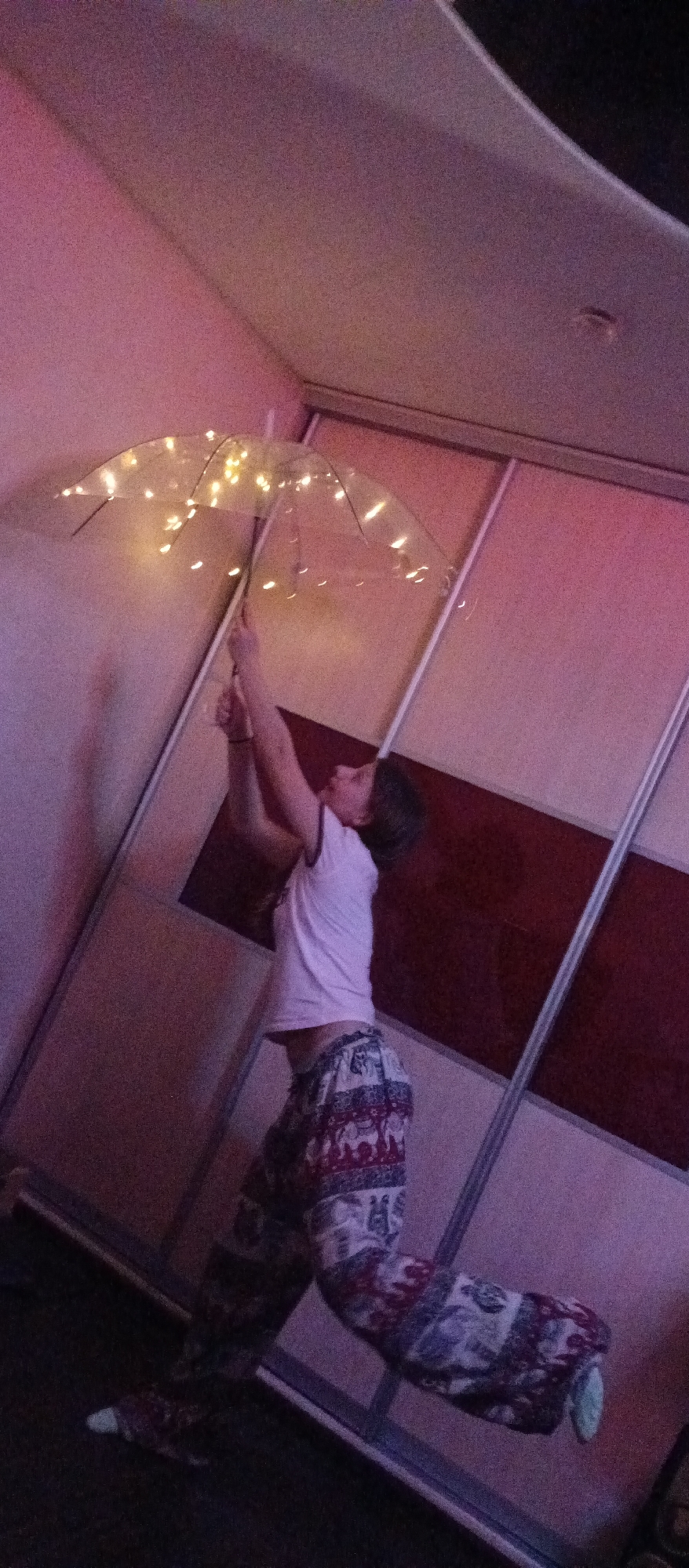 An indoor scene captures a person, likely a child or young adult, holding a clear umbrella adorned with illuminated string lights. The individual wears a plain white t-shirt and patterned trousers, which feature repetitive elephant motifs. The person is actively holding the umbrella upwards with both hands, gazing at it, and positioned with their body angled and their left leg bent at the knee, suggesting a playful leap, dance, or an attempt to "float."
In the background, there is a wardrobe or closet with sliding doors, some panels appearing as a dark reddish-brown and others in lighter tones. The room is dimly lit, with a distinct purple and pink ambient glow, implying an evening or nighttime setting. The illuminated string lights on the umbrella further contribute to this atmospheric lighting. The overall impression is one of imaginative playfulness and whimsy within a domestic environment. No text is visible in the image.
