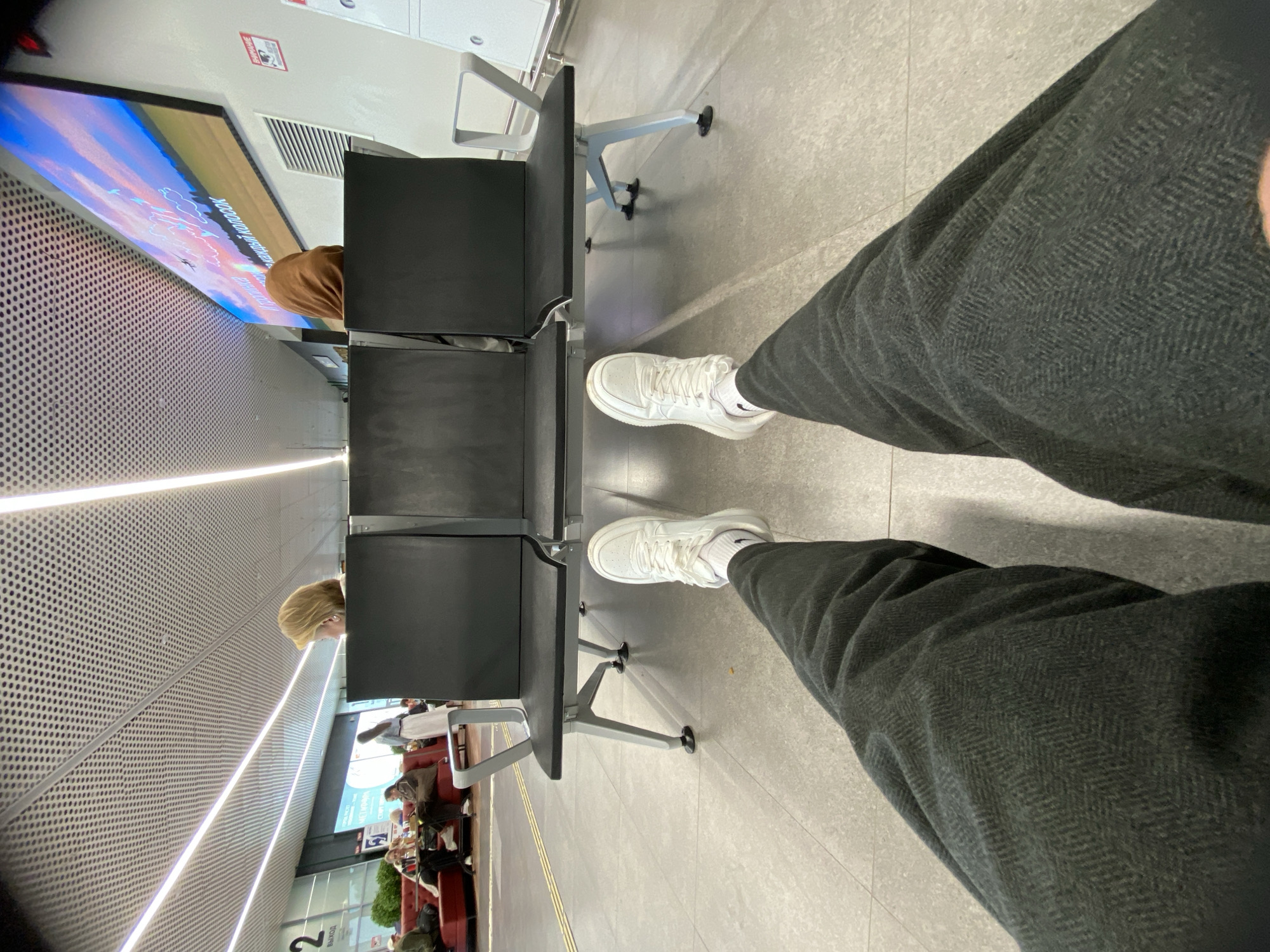 The image shows a first-person perspective of a person sitting in an airport waiting area. The person is wearing dark herringbone-patterned pants and white sneakers. They are seated in a row of black airport chairs, with their legs extended forward. In the background, other passengers are visible sitting on red couches and walking. A large screen displays an image of a sky with clouds and an airplane. The location is identified as an airport in Новое Мартьяново, Russia, indicated by Cyrillic text on signage in the background. The environment appears clean and modern, with a perforated ceiling and bright lighting.