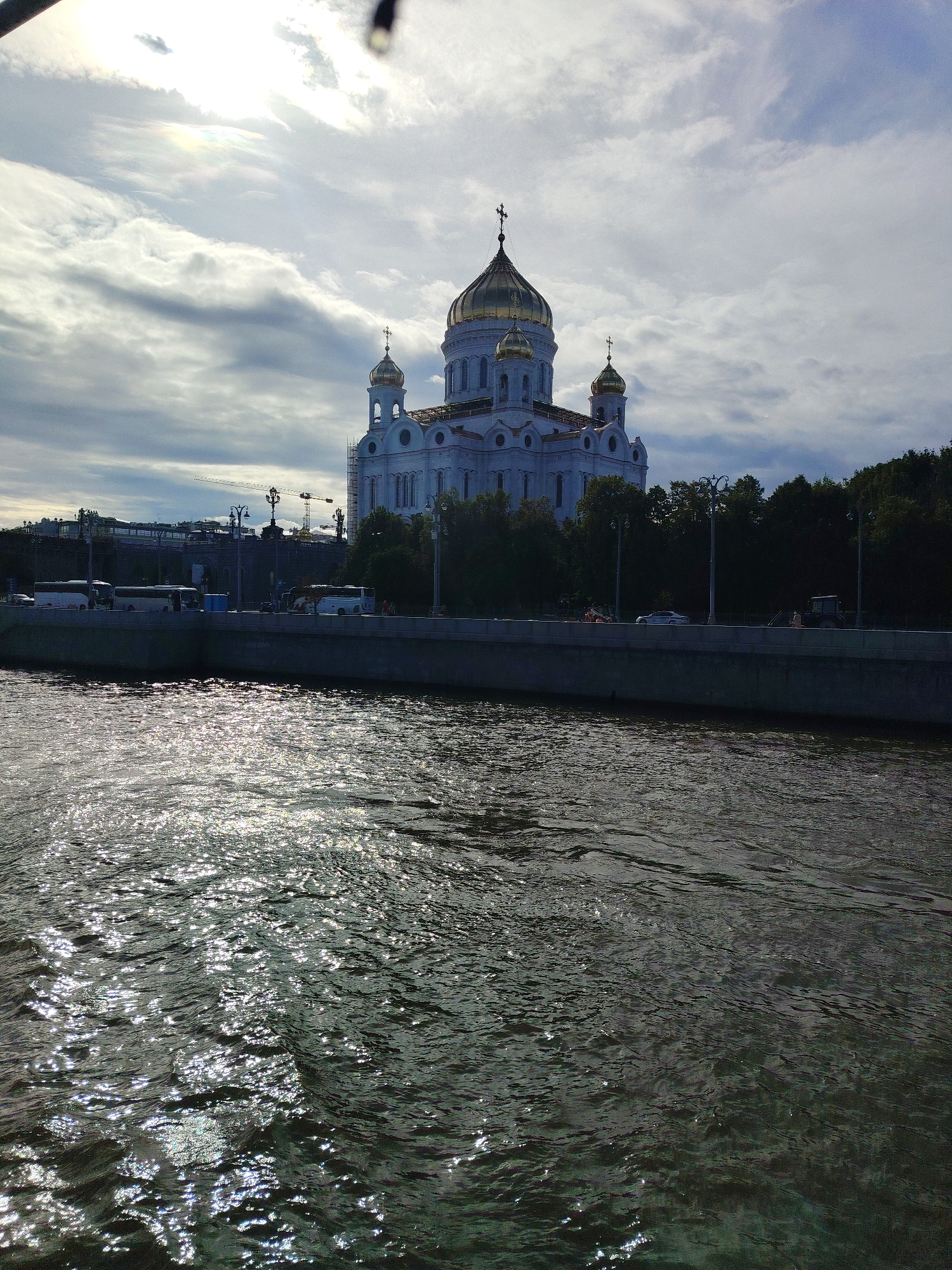 The image depicts a wide, dark river in the foreground, with its surface shimmering with sunlight reflecting off the ripples. A stone embankment runs along the river's edge. Beyond the embankment, a white building with golden domes, likely a church, stands prominently. Trees line the area behind the building, and in the distance, there are indications of urban infrastructure, including what appear to be buses parked on a street or promenade and cranes, suggesting ongoing construction. The sky above is cloudy with patches of blue and sunlight breaking through.
