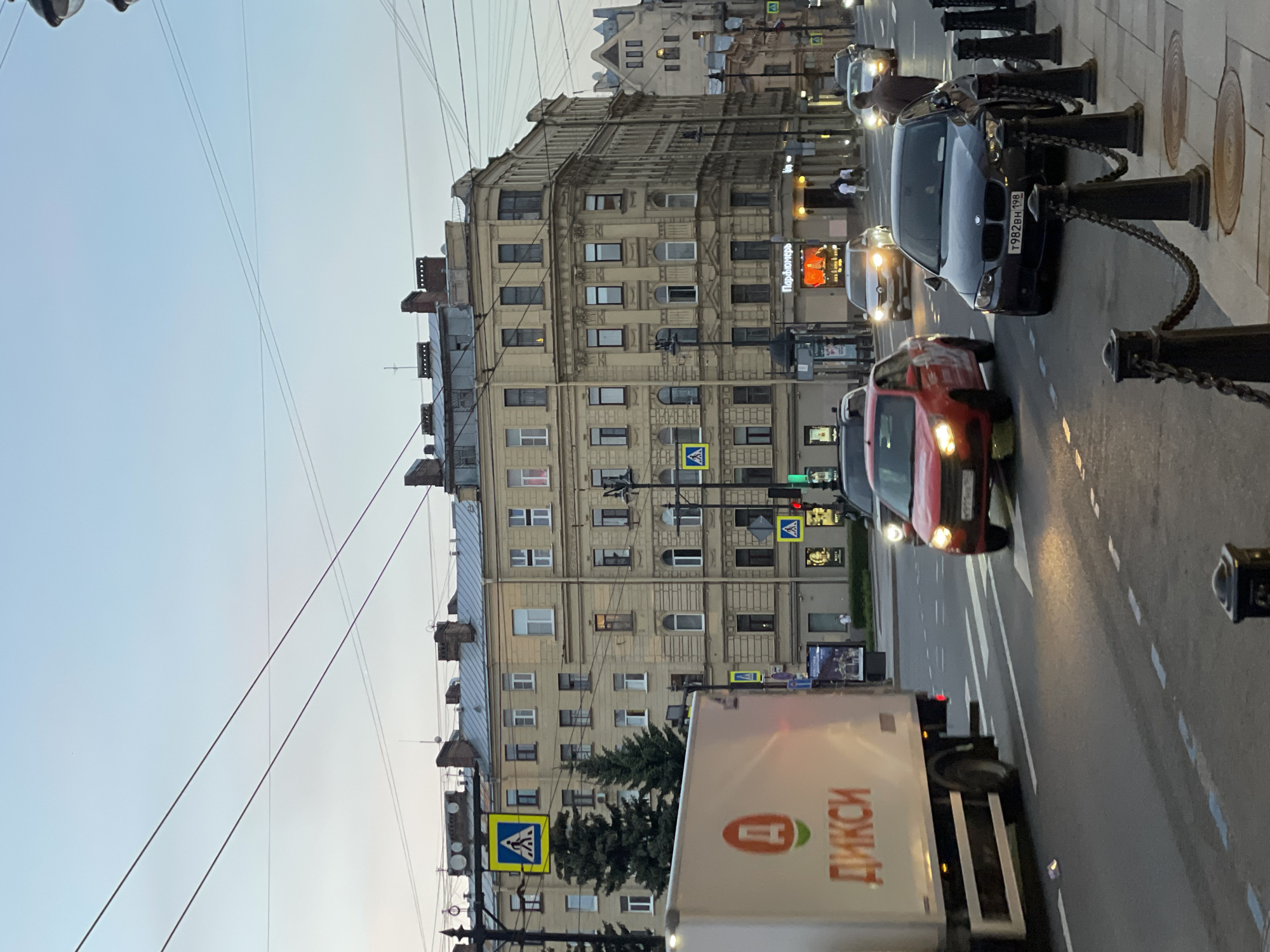 The image shows a street in Saint Petersburg, Russia. A large beige building with multiple windows and architectural details dominates the left side. Overhead wires crisscross the sky. Street-level elements include pedestrian crossing signs, traffic lights, a white truck with Cyrillic script reading "Дикси" (Dixy), a red car, a black BMW with license plate "T982BH 198", and black bollards with chains lining the sidewalk. Other cars are visible in the distance. The sky is pale blue, suggesting either early morning or late evening.