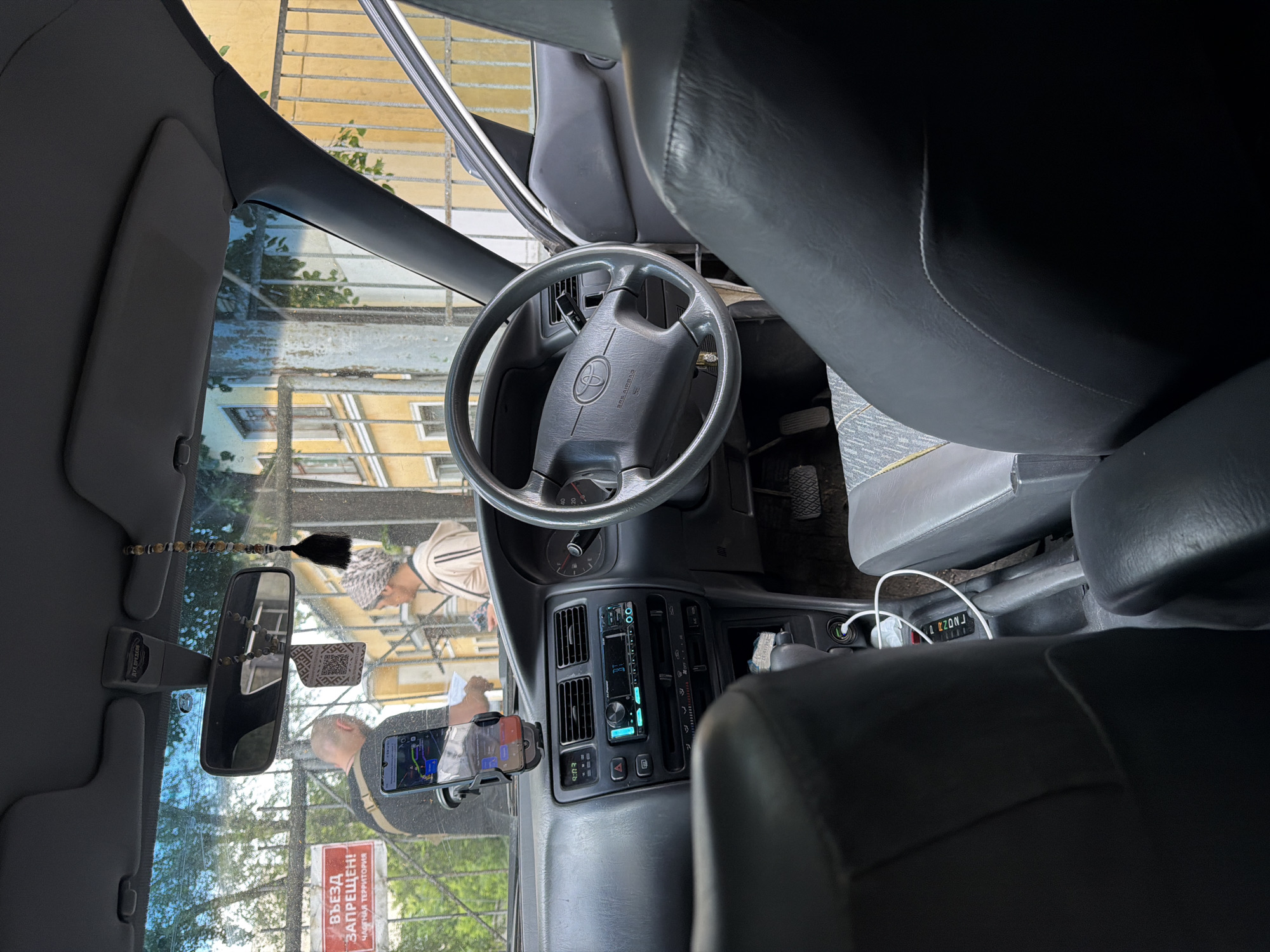 The image shows the interior of a car, likely a right-hand drive vehicle, looking out through the windshield. Outside, a man and a woman are standing near a metal fence. The man is wearing a dark t-shirt and has a crossbody bag. A smartphone is mounted on the dashboard, displaying a navigation app. The woman is wearing a light-colored hoodie and a headscarf. Behind them, a yellow building with windows is visible. A red sign on the fence reads "ВЪЕЗД ЗАПРЕЩЕН! ЧАСТНАЯ ТЕРРИТОРИЯ," which translates to "ENTRY PROHIBITED! PRIVATE TERRITORY." The car's interior features a dashboard with air vents, a car stereo, and a steering wheel with a Toyota logo. The seats are upholstered in dark material with some patterned accents on the passenger seat. The overall scene suggests a moment captured at the entrance to a private area in Moscow, Russia, where the individuals are either being denied entry or are discussing their access.