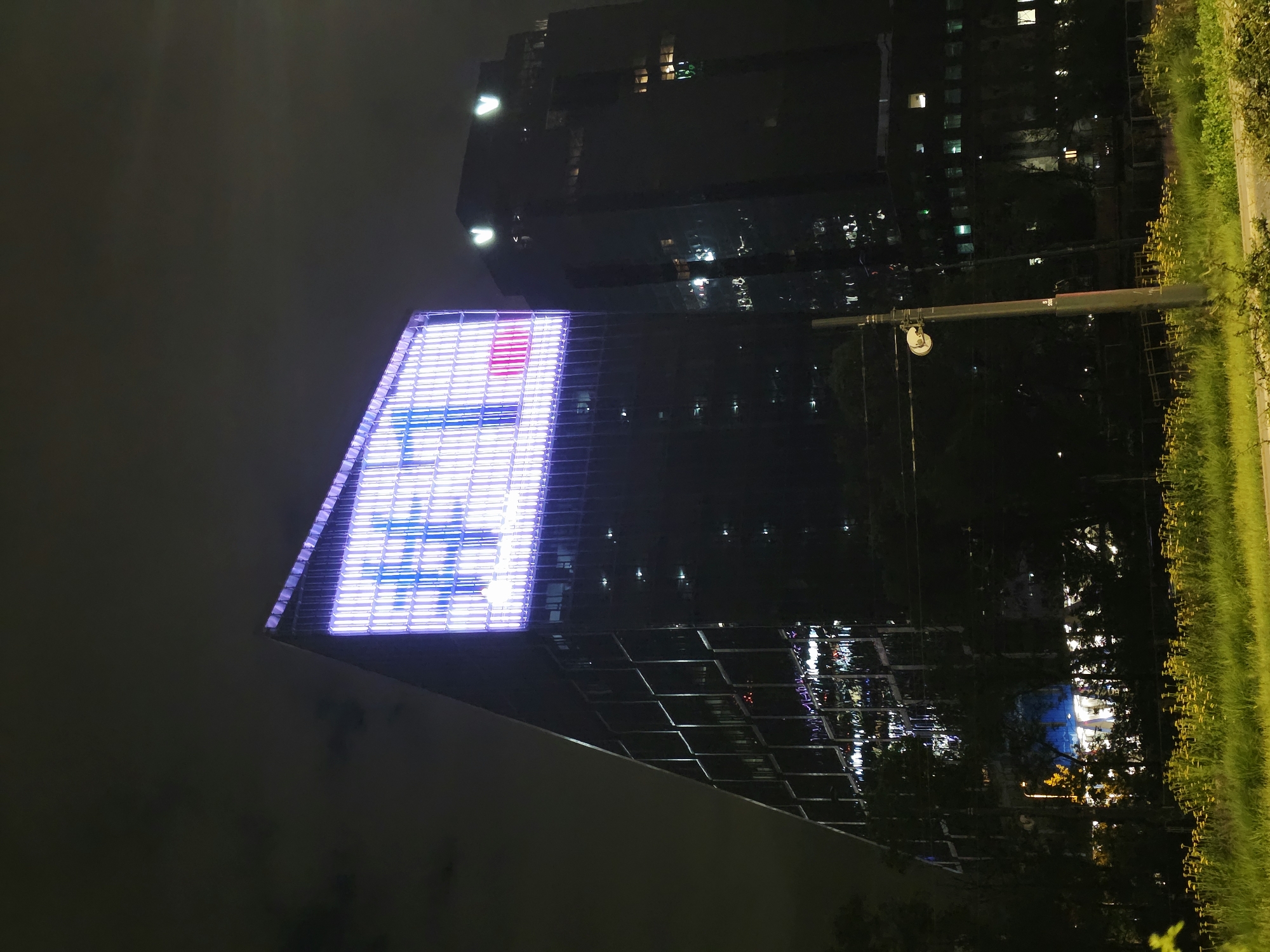 The image depicts a night scene in Warsaw, Poland, featuring two modern high-rise buildings. The building on the left is angled and illuminated by a large, colorful digital display that appears to show a grid of lights with some blue and pink elements. The building on the right is darker and also has illuminated windows. In the foreground, there is a grassy area with yellow flowers and a lamppost. Trees and other vegetation are visible between the foreground and the buildings. The sky is dark and cloudy.