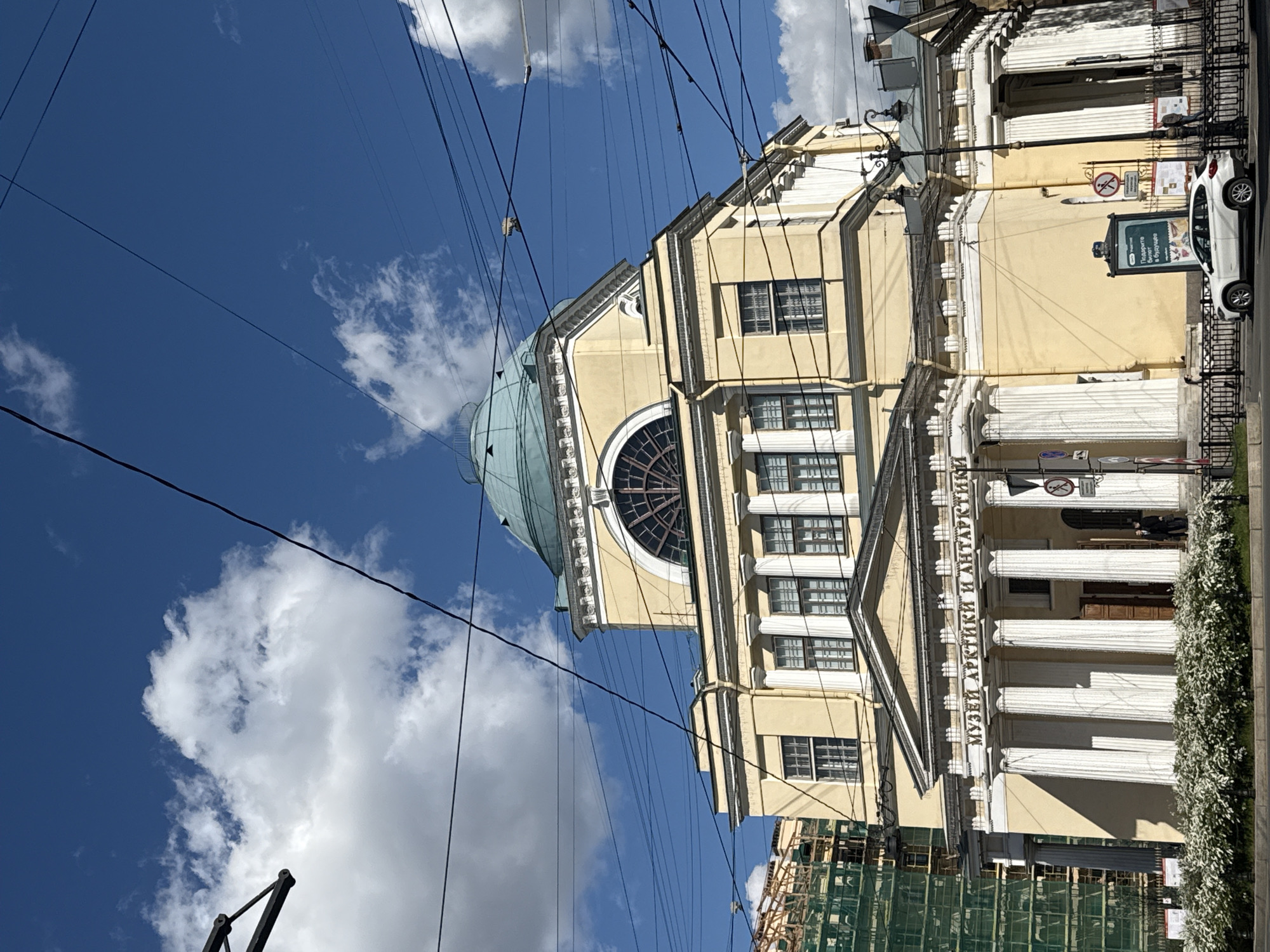 The image depicts the Russian State Museum of the Arctic and Antarctic in Saint Petersburg, Russia. The building is cream-colored with a distinctive dome and a facade adorned with classical columns. The museum's name is inscribed in Russian along the facade. Above the building, power lines are visible against a blue sky with scattered clouds. A white car is parked to the right of the building along a street lined with greenery. Scaffolding partially obstructs the view of the lower part of the building. A sign is displayed in front of the museum. The orientation of the image is rotated counter-clockwise.
