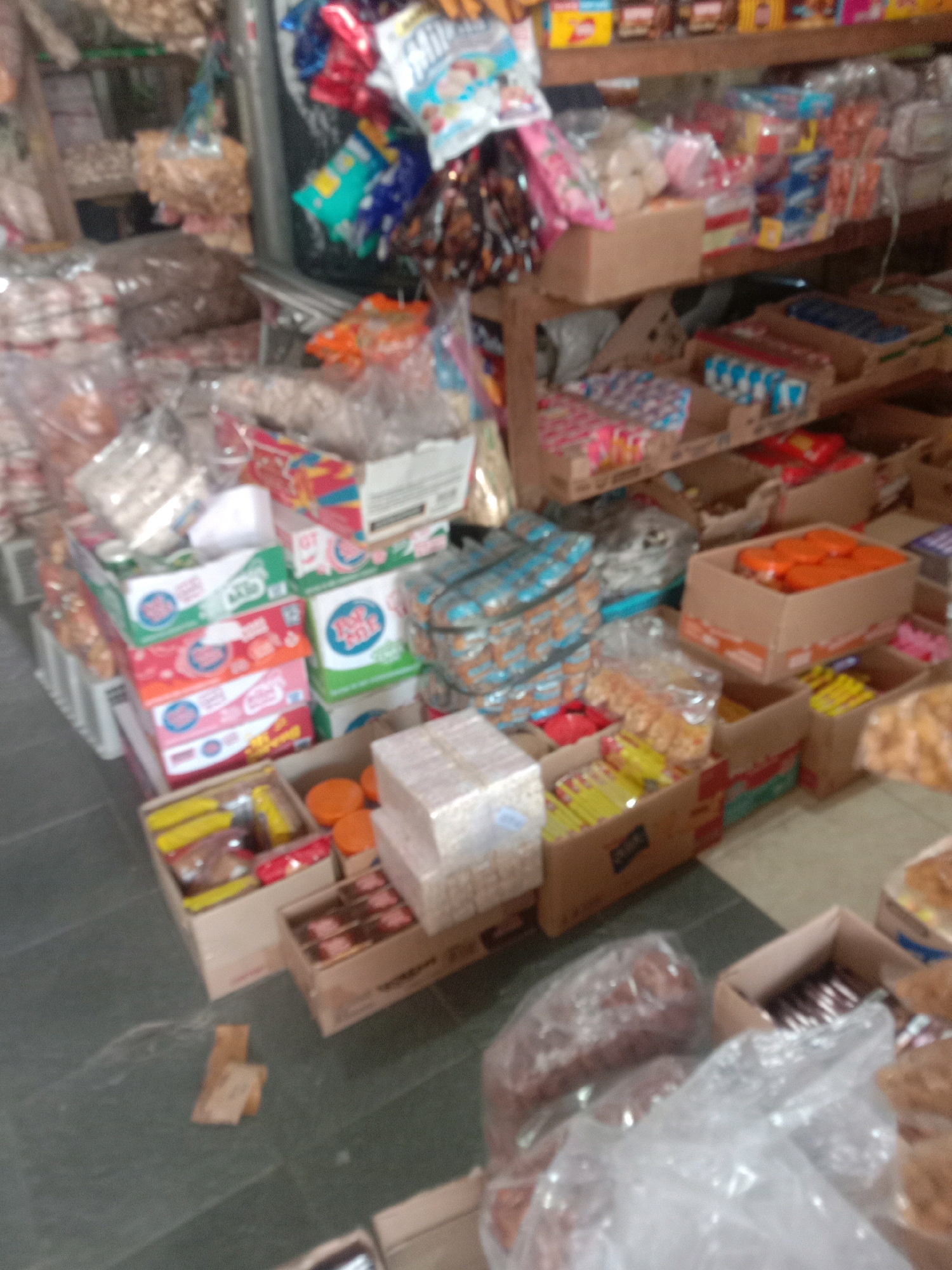 The image depicts a cluttered market stall in Sidogiri, Indonesia, overflowing with various packaged snacks and goods. Rows of brightly colored boxes, many with visible text and graphics, are stacked haphazardly on shelves and on the floor. Some of the boxes appear to be food items like crackers or biscuits, indicated by the packaging. There are also bags of what seem to be dry goods, possibly nuts or snacks, displayed in clear plastic packaging. In the foreground, a large clear plastic bag contains a substantial amount of brown, loaf-like items, possibly bread or baked goods. The overall impression is one of a busy, small-scale retail environment where a wide variety of products are densely packed for sale. The background shows more shelves lined with similar merchandise, suggesting a limited space with a high volume of inventory.