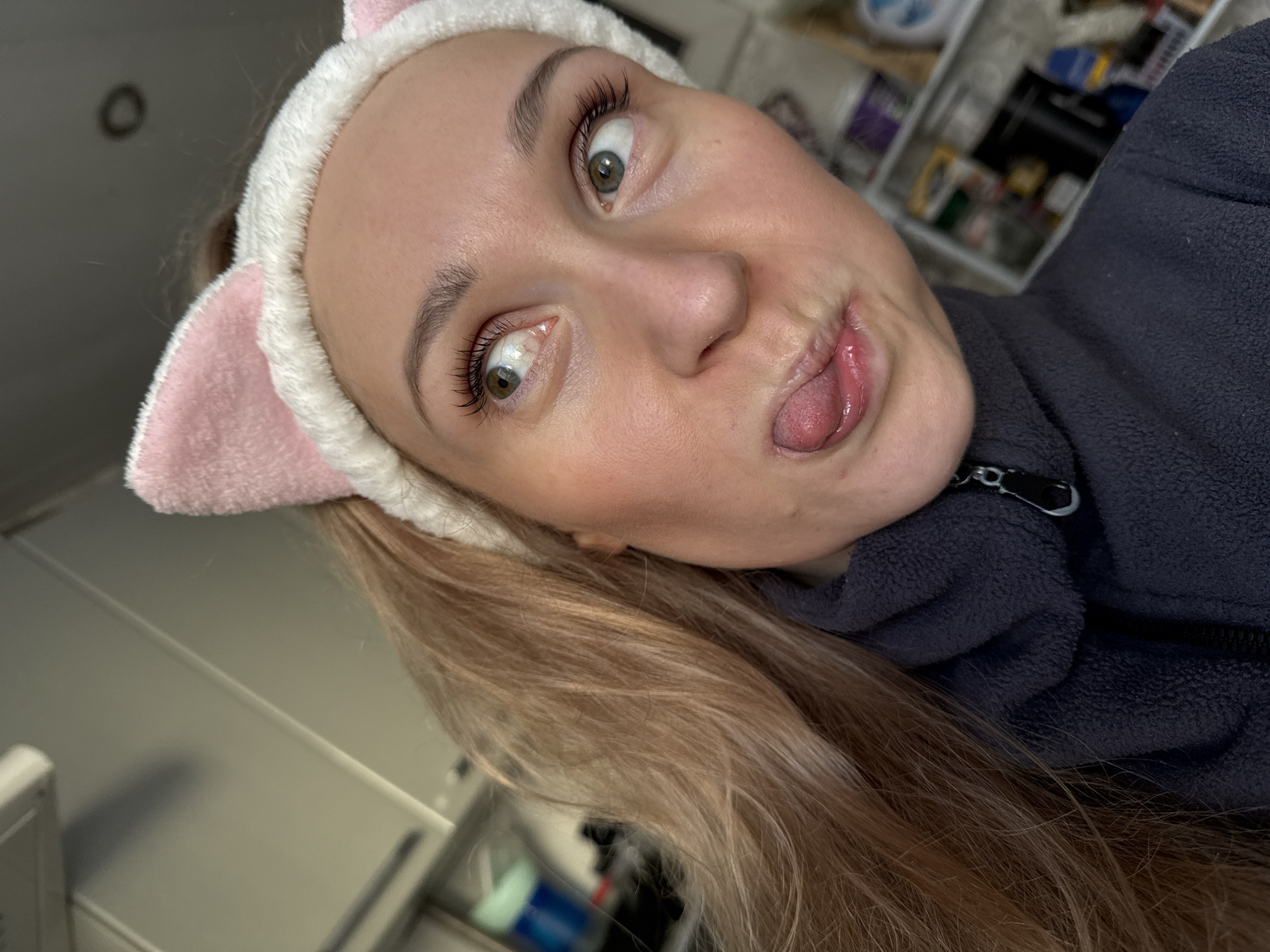 A selfie of a fair-skinned woman wearing a pink and white plush cat-ear headband, sticking her tongue out, and wearing a dark zippered jacket. Her hair is light brown and partially obscures the bottom left of the frame. The background includes a light-colored cabinet and miscellaneous items on shelves. The location is not discernible from the image.