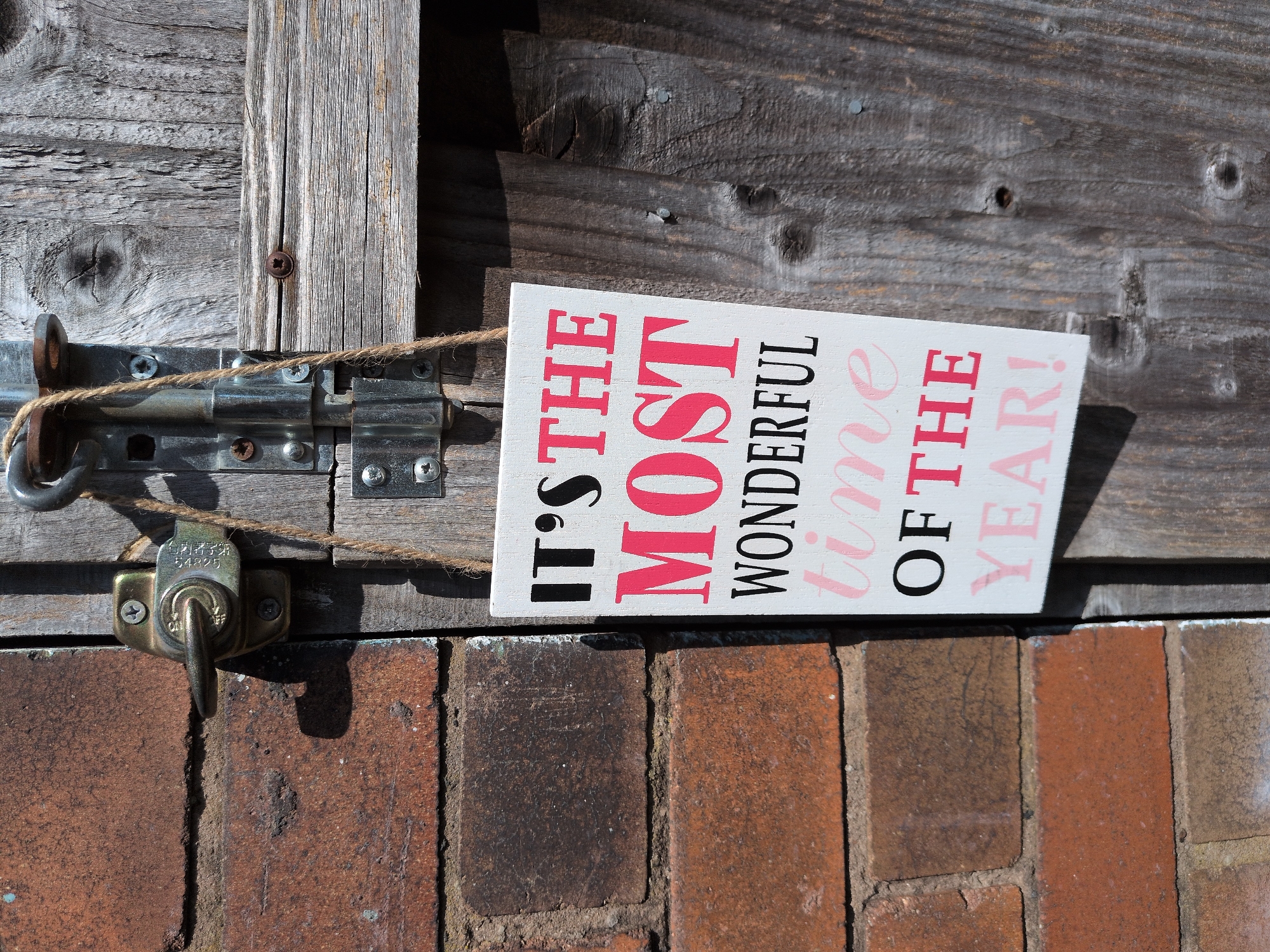 A white wooden sign that reads "IT'S THE MOST WONDERFUL TIME OF THE YEAR!" in black and pink lettering is hanging from a metal latch on a weathered wooden gate. The gate is situated next to a brick wall. The scene is outdoors with sunlight casting shadows. The sign is attached to the gate by a piece of twine. The latch appears to be a sliding bolt mechanism. The bricks are a reddish-brown color, and the wood of the gate is gray and shows signs of age, with visible grain and some cracking. The image is taken from a slightly high angle, looking down at the sign and gate.