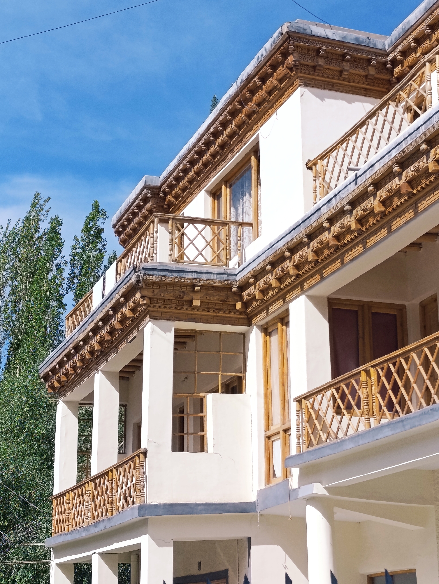 The image shows the exterior of a traditional Ladakhi building in Leh, India. The building features white stucco walls and intricately carved wooden balconies and eaves. The balconies have a lattice-like railing design, and the eaves are adorned with detailed geometric and floral carvings. Tall, slender trees with green foliage are visible to the left of the building, and a clear blue sky with a few wispy clouds is overhead. The architectural style suggests it is a guesthouse or residential building, typical of the region. The overall impression is one of a serene and culturally rich environment.