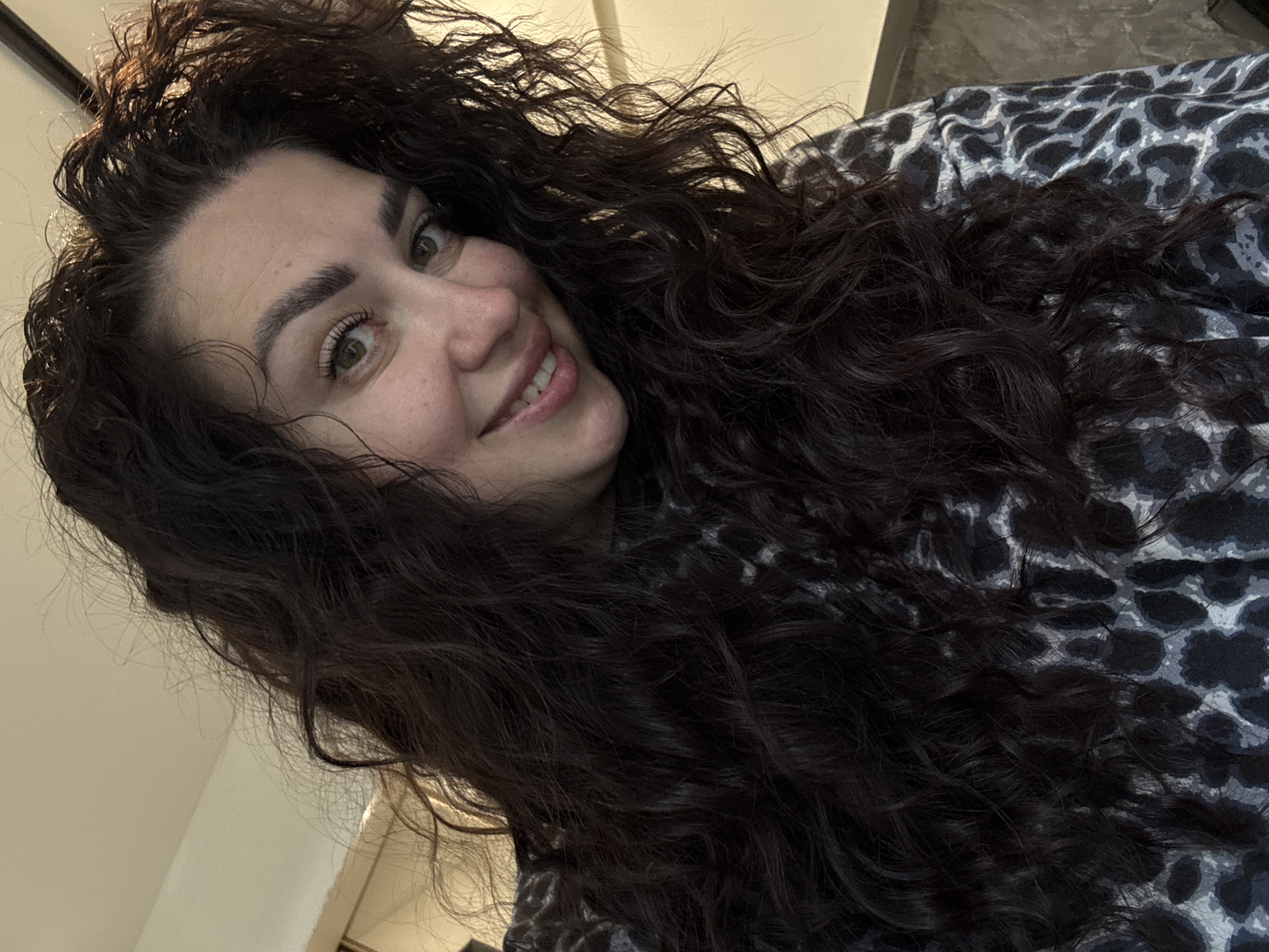 A close-up, high-angle selfie shows a woman with long, dark, curly hair and light green eyes. She has a warm smile and is looking towards the camera. The woman is wearing a grey and black animal print top. The background consists of a beige wall and a dark brown surface, suggesting an indoor setting. The lighting is soft and appears to be coming from the right. The overall impression is informal and relaxed.