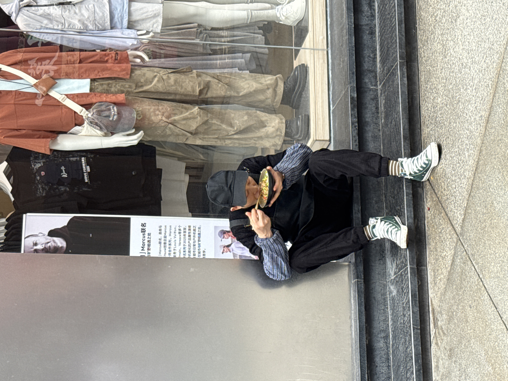 A man is squatting on the sidewalk in Jinjiang District, China, eating from a bowl with chopsticks. He wears a black cap, a black jacket over a striped long-sleeve shirt, and dark pants. He is wearing green and white sneakers with brown socks. He sits next to a shop window displaying mannequins dressed in various clothes, including pants, jackets, and shirts. The shop window also has a poster with the words "Marcus联名 与旷野相遇之处" and "MOVE FORWARD with friends" written on it. The sidewalk is made of grey concrete. The photo is taken from a high angle.
