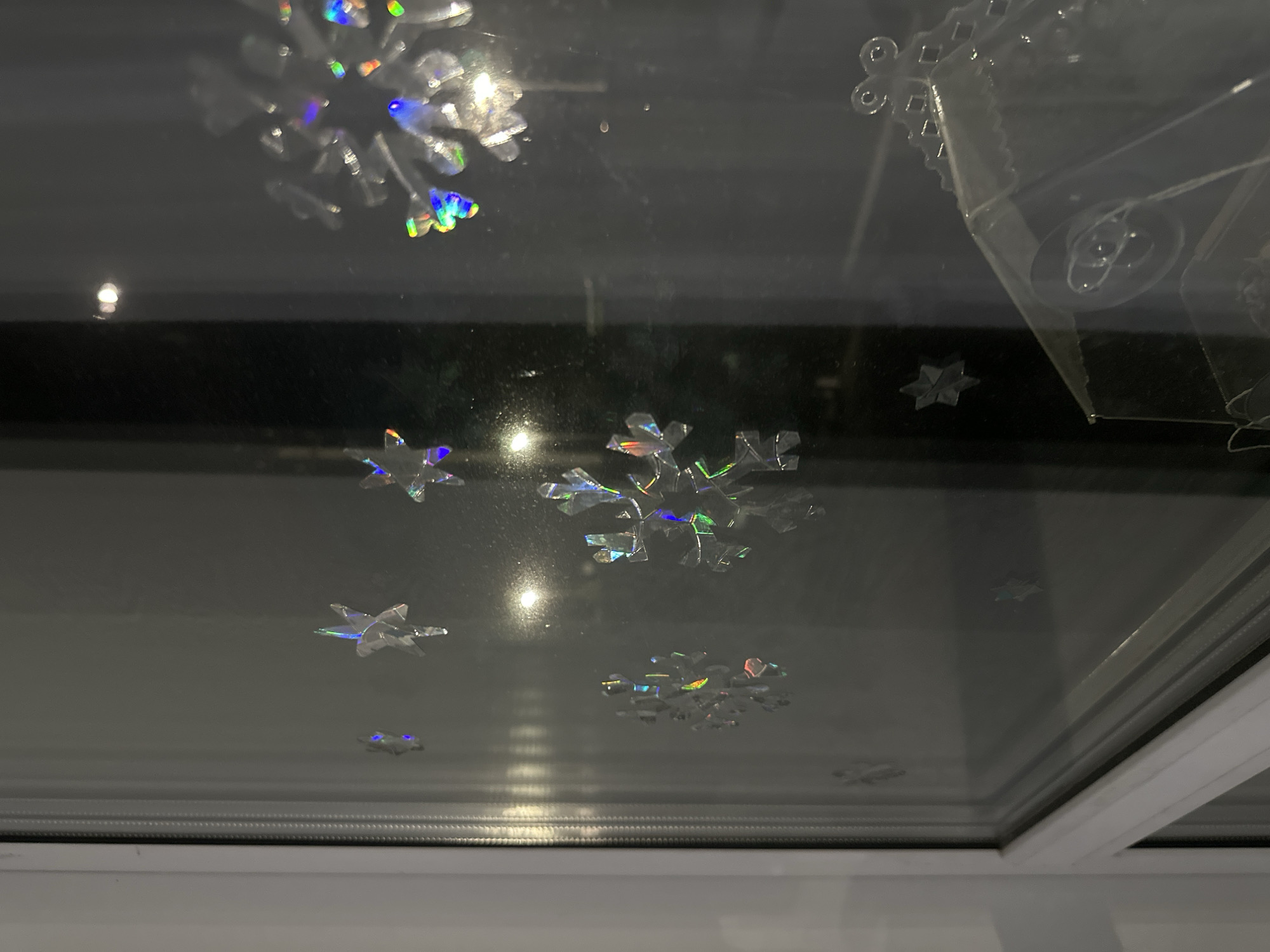 The image shows a window adorned with snowflake-shaped stickers. The stickers appear to be holographic, reflecting light in various colors. The window frame is white. The location is Unknown, Unknown.