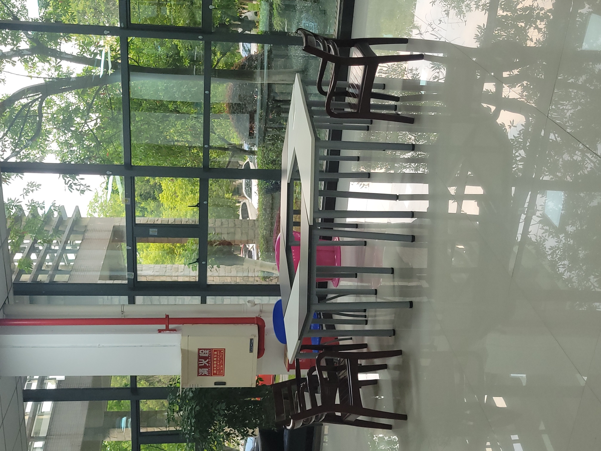 The photograph, taken in Chongqing High-tech Zone, China, depicts an indoor space with large windows. Outside, trees and vegetation are visible, and their reflections are prominent on the shiny tiled floor.
Inside, there is a long, white table with metal legs. Several chairs, both wooden and plastic, are arranged around the table. A fire extinguisher and a red pipe are mounted on the wall near the table. The lighting is bright, likely due to the large windows and reflective floor. The space appears to be a cafeteria or a meeting room.