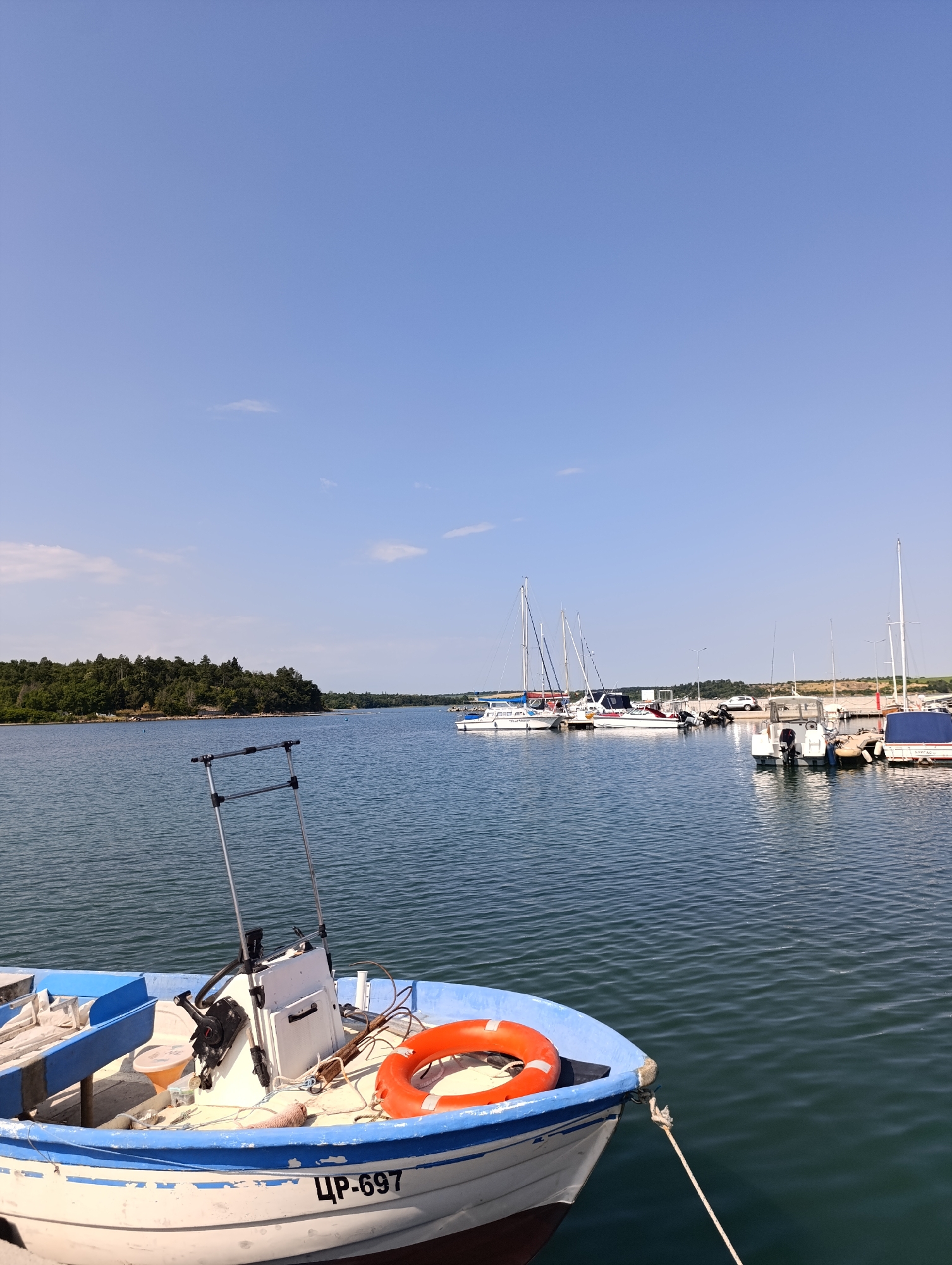 A medium shot captures a bright, sunny day at a marina in Burgas, Bulgaria. In the foreground, a white and blue fishing boat is docked, with its registration number "ЦР-697" clearly visible on the hull. An orange life preserver is on deck, along with a steering console and fishing equipment. The water is calm, with a gentle ripple. In the background, several sailboats and motorboats are moored. A lush, green forest lines the shore of an island in the distance. The sky is a clear, pale blue with a few wispy clouds. The scene depicts a typical day at a coastal Bulgarian port.