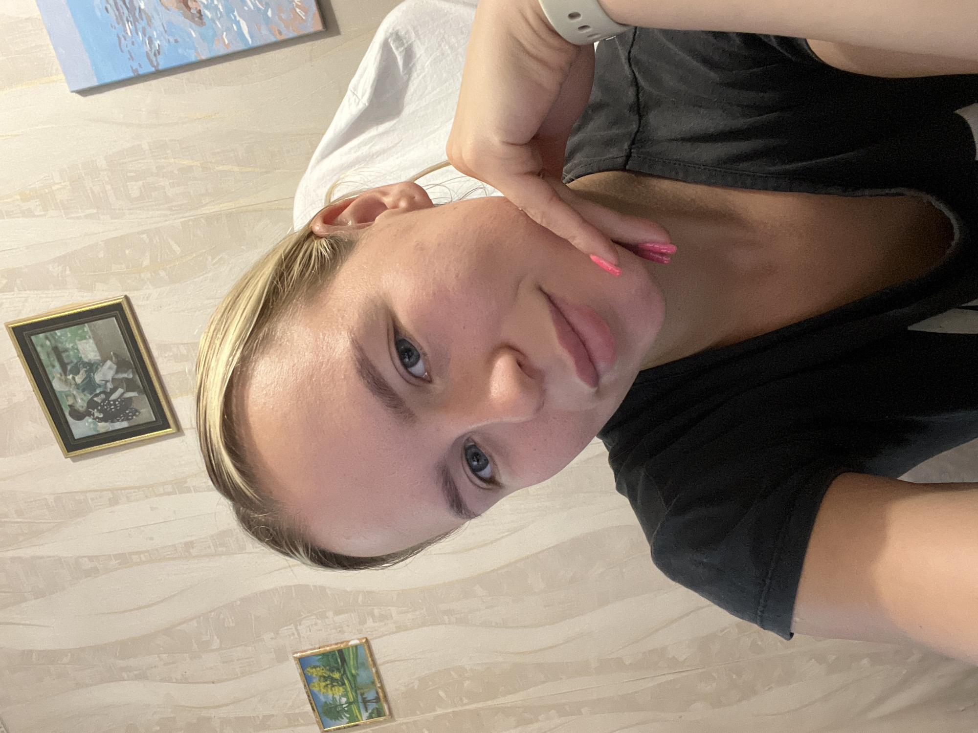 The media file is a selfie of a young woman with blonde hair. She is wearing a black t-shirt. Her left hand is touching her face. She has bright pink nail polish on her fingernails. She is lying down, as indicated by the white fabric behind her head, which appears to be a pillow or bedding. The background wall has a wavy pattern and two small framed pictures hang on it. One picture is dark, and the other appears to be a landscape. The lighting suggests it might be daytime, but the specific time of day is not discernible. The woman's expression is neutral. There is no visible text in the media file.