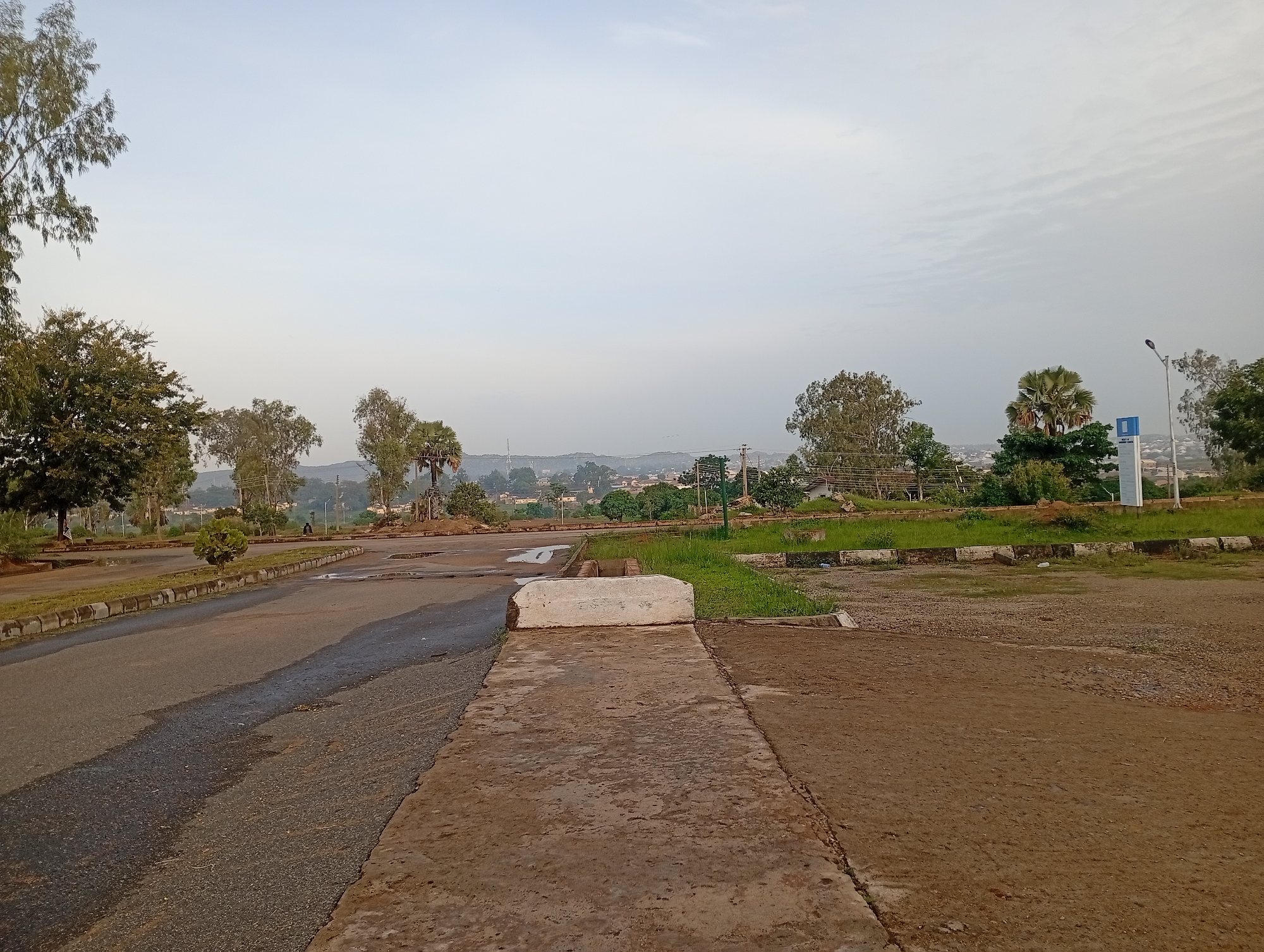 The image depicts an outdoor scene, likely in the city of Jos, Nigeria, during what appears to be early morning or late afternoon, given the soft lighting. In the foreground, a paved road runs diagonally from the left to the center of the frame, showing patches of wetness and shadows. To the right of the road is a dirt area with scattered gravel. A concrete pathway extends into the scene from the bottom center, leading towards a white rectangular object, possibly a bench or a bollard, positioned on the edge of the pathway.
Beyond the immediate foreground, a grassy area with some scattered trees and shrubs is visible. In the middle ground, a wider paved area, possibly a parking lot or open space, contains some puddles of water. Further in the background, a town or city landscape is visible with buildings and trees, suggesting an urban or semi-urban environment. Several palm trees are prominent in the mid-background. The sky is overcast with a pale, diffused light. A single street lamp is visible on the right side of the image, near a blue and white sign. A lone figure is seated on the left side of the paved area in the mid-ground. The overall impression is one of an undeveloped or somewhat neglected public space.
