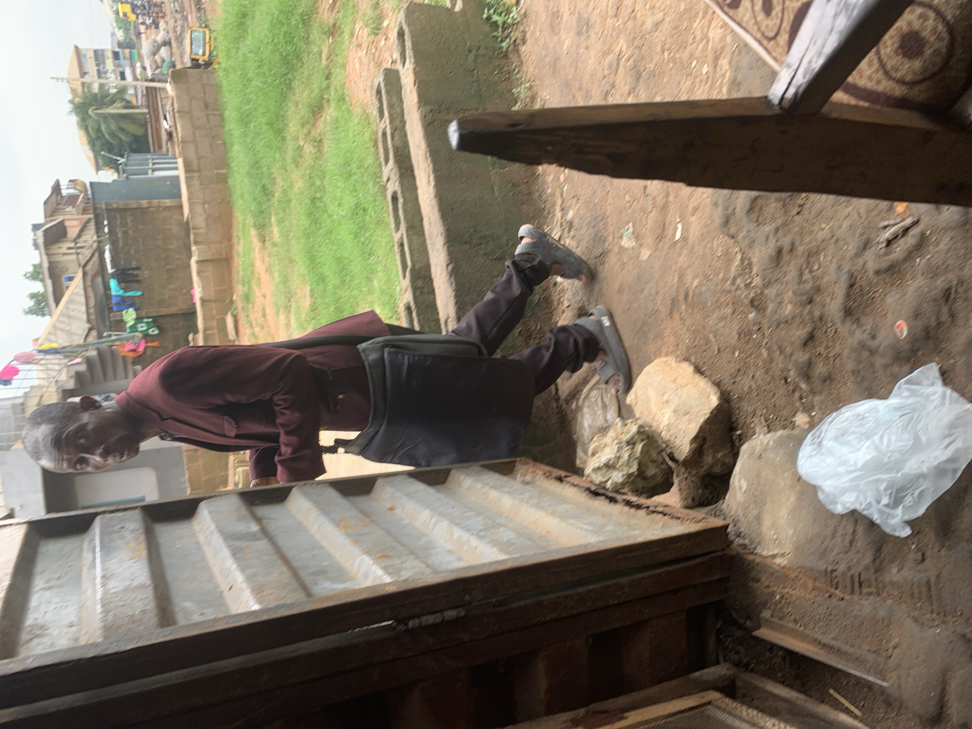 A man wearing a maroon jacket and dark pants walks past an open, corrugated metal door in Shagamu, Nigeria. He carries a black bag on his left shoulder and is wearing grey sandals. The ground is dirt with scattered rocks. Behind the man, there are concrete blocks and green grass. In the background, buildings, laundry hanging on a line, and a yellow vehicle are visible. The sky is overcast.