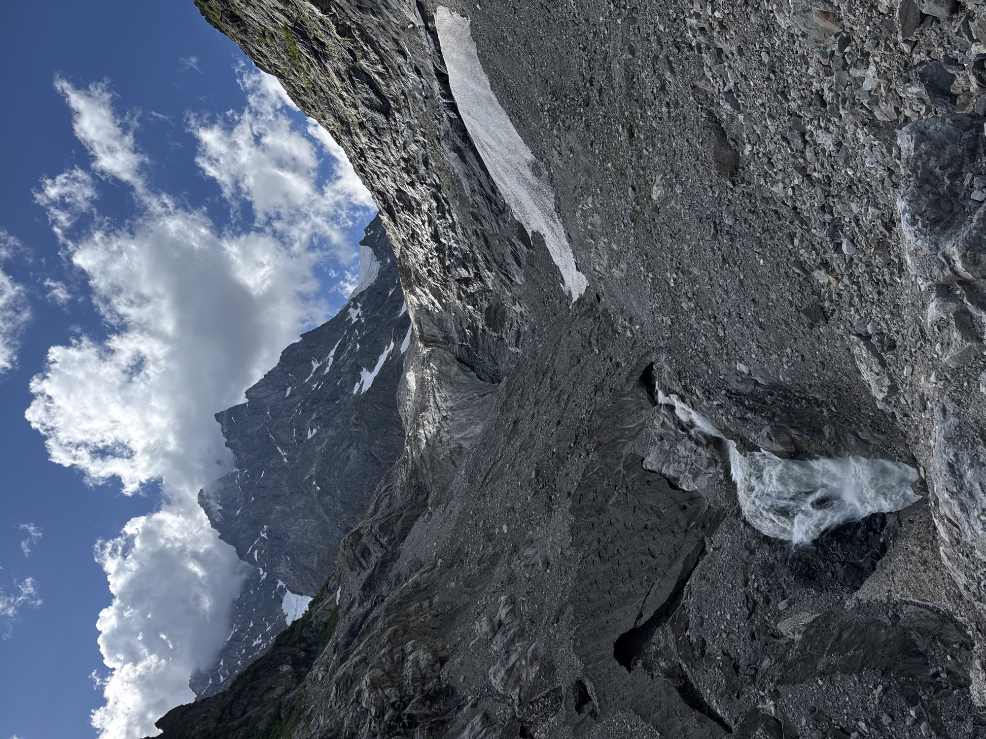 The image depicts a dramatic, high-altitude mountain landscape. In the foreground, a powerful waterfall cascades down a steep, rocky slope covered in scree and debris. The water is white and frothy, indicating a rapid descent. Above the waterfall, the terrain is characterized by rugged, dark gray rock formations interspersed with patches of snow and ice. A glacier is visible further up the valley, with its textured surface reflecting sunlight. The surrounding mountains are jagged and imposing, with sharp peaks piercing a bright blue sky dotted with fluffy white clouds. Some of these peaks are capped with snow. The overall environment is remote and wild, suggesting a glacial valley in a mountainous region. The image does not contain any people, structures, or any other elements that would identify a specific location within the city of Murshkeli, Georgia.