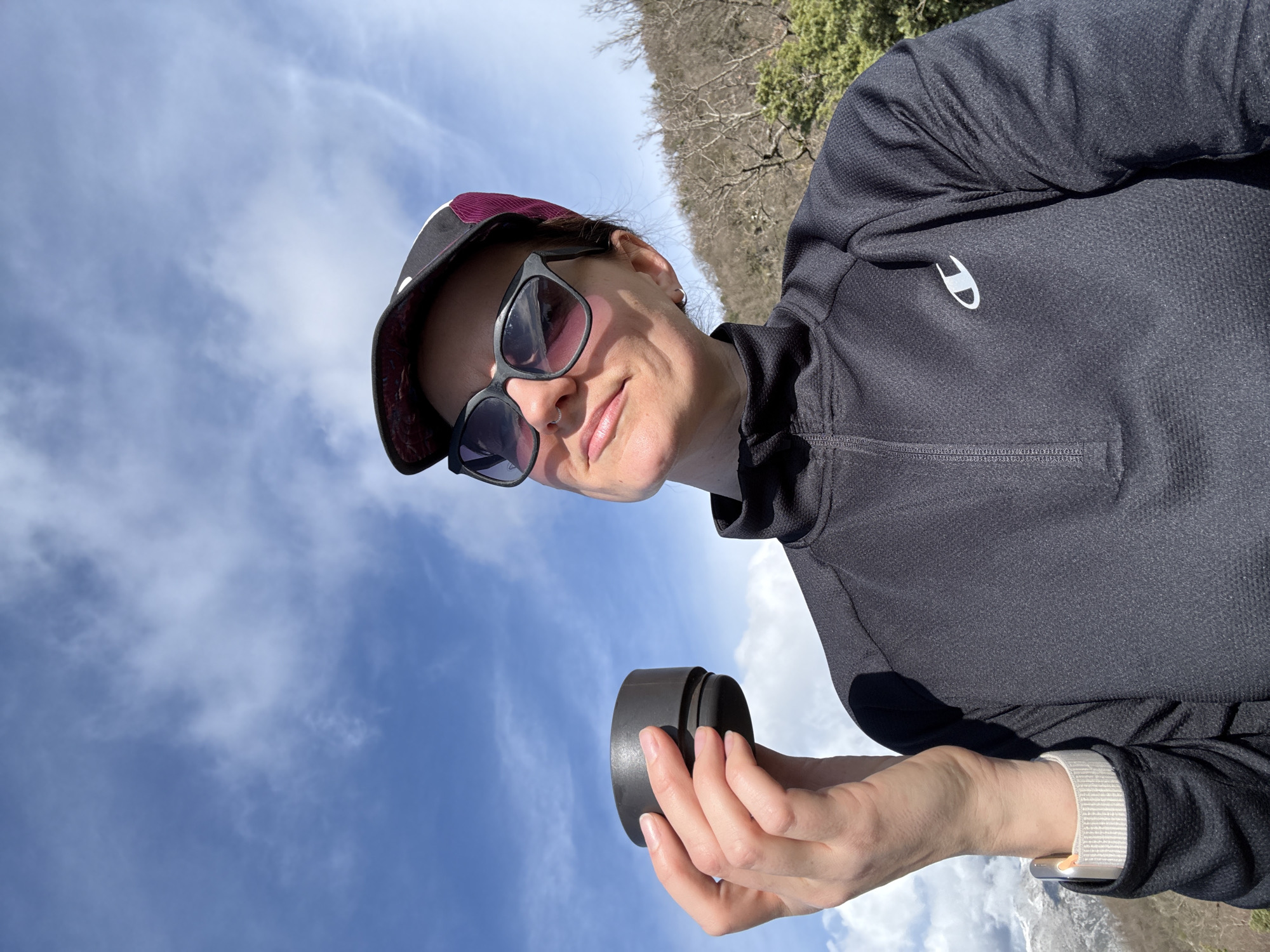 An outdoor image captured in Gornja Lastva, Montenegro, depicts a female subject from the chest up, positioned against a sky and natural landscape background. The subject wears a dark-colored baseball cap with a white front panel and a patterned brim, along with dark-framed sunglasses. A small nose ring is visible on her left nostril, and a silver hoop earring is in her right ear. She is dressed in a black quarter-zip athletic shirt, featuring a white Champion logo on the left chest. Her right hand holds a dark grey or black cylindrical cup. A light-colored smartwatch with a woven band is visible on her left wrist. The subject is looking upwards and slightly to her right. The background features a clear blue sky with scattered white clouds. To the right, a hillside with bare deciduous trees and some green foliage is present. In the lower left background, distant snow-capped mountains are partially visible.