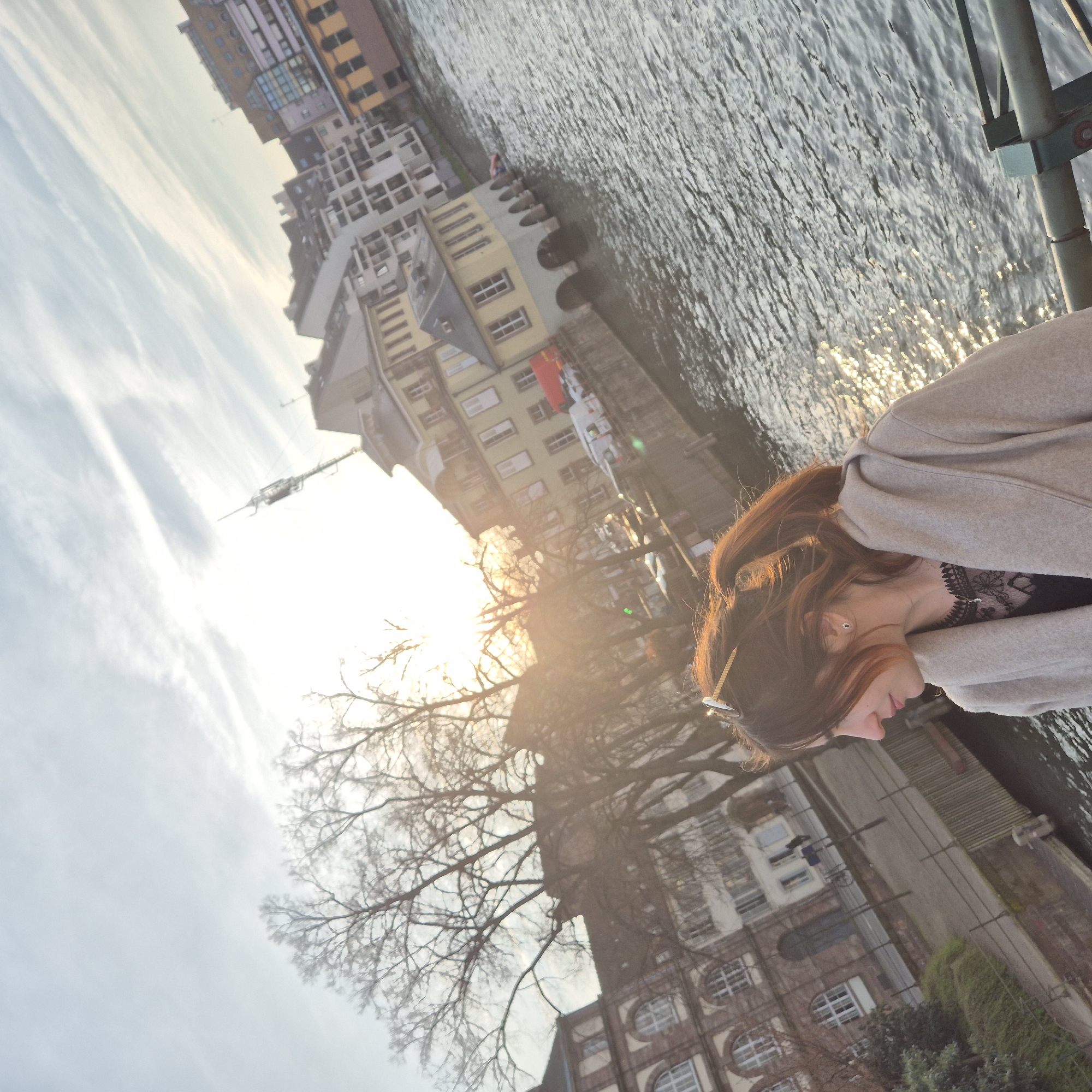 The image depicts an outdoor urban scene in Strasbourg, France, specifically along a river or canal. The perspective is slightly tilted, with the sun low in the sky, causing significant lens flare in the upper left quadrant.
In the foreground, positioned on the right, a female individual is captured in profile, looking towards the left. She has reddish-brown hair tied back in a ponytail, with sunglasses resting on top of her head. She is wearing a light beige or tan hooded coat over a dark top, possibly black, with visible lace detailing at the neckline. A delicate necklace with a small pendant and a stud earring are discernible. A green and grey metal railing, consistent with a bridge or embankment barrier, is present in the bottom right corner.
The mid-ground features a body of water, likely the Ill River, exhibiting ripples and reflecting bright sunlight. On the opposite bank, a collection of historic buildings lines the waterfront. One prominent yellow-painted building features multiple arches at its base, extending directly into the water, suggesting a boat-house or a structure built directly on the river. To its left, another building displays a reddish-brown brick facade with large arched windows. Bare deciduous trees, devoid of leaves, stand along the riverbank. A street or pedestrian path runs parallel to the river on the far bank, with several parked vehicles, including at least one red van and multiple white vans. A street lamp and a bicycle are also visible on this bank.
The background shows additional city buildings of varying architectural styles and heights. Against the bright sky on the upper right, a tall, slender antenna or communication mast is visible. The sky is bright with scattered, thin clouds. The overall lighting conditions suggest late afternoon or early morning, with the bare trees indicating either a late autumn or winter setting. No specific actions or interactions are depicted beyond the individual in the foreground posing for the photograph.