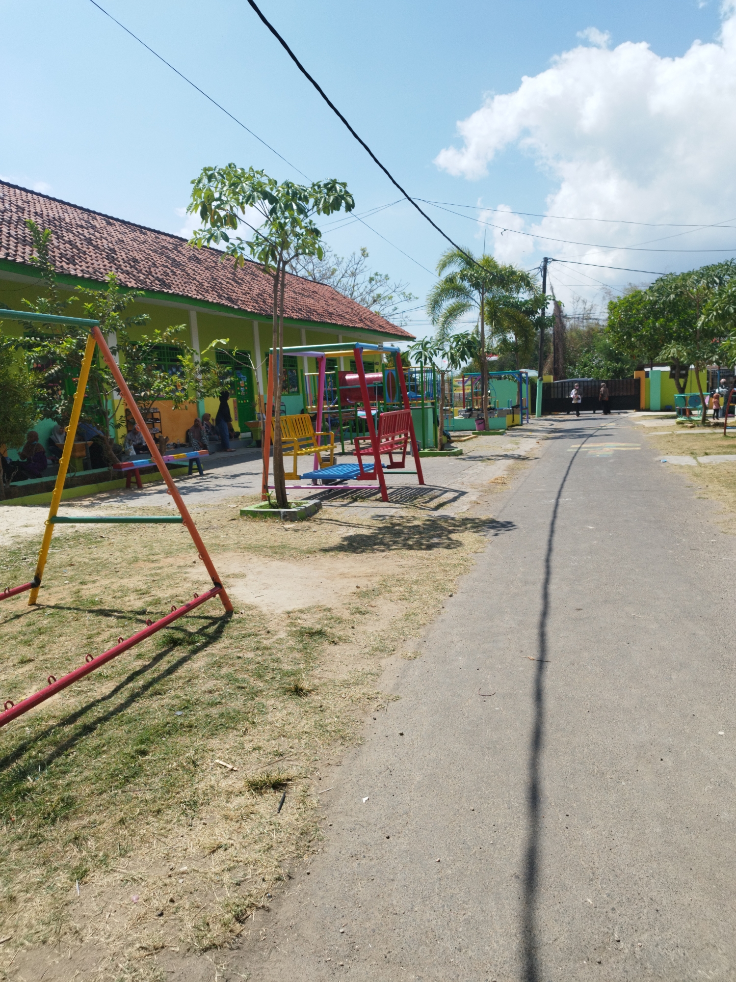 The image depicts an outdoor scene at what appears to be a playground or school area in Marengan Daya, Indonesia. A paved pathway runs through the center of the image, with a grassy and dirt area on the left. To the left of the pathway is a colorful playground with swings, a seesaw, and other play structures. A one-story building with a red tile roof and light green walls is visible behind the playground. Several people, including children and adults, are present in the area, some sitting on benches and others walking. The sky is blue with scattered clouds. The overall environment is bright and sunny, suggesting a warm day.
