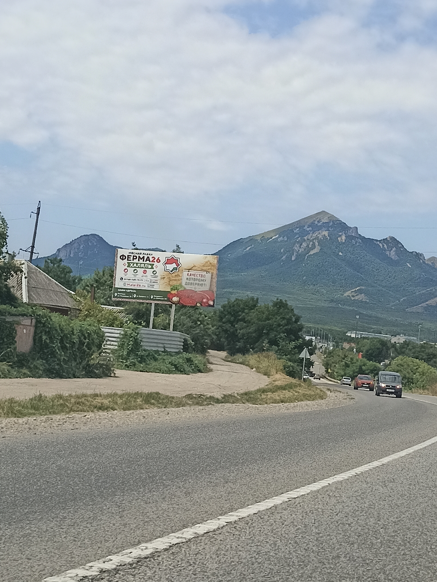 The image captures a rural or suburban roadside scene with a prominent mountain range in the background. A billboard for "FERMA26" is visible on the left, displaying images of produce and meat, along with text in Cyrillic. In front of the billboard is a small, rustic building surrounded by dense green foliage. The road in the foreground curves to the right, with several vehicles, including a van and a car, driving in the same direction. The sky is overcast with scattered clouds. The location is identified as Inozemtsevo, Russia, a town known for its proximity to the Caucasian Mountains. The overall impression is one of a roadside view on a cloudy day in a scenic, possibly agricultural, region.