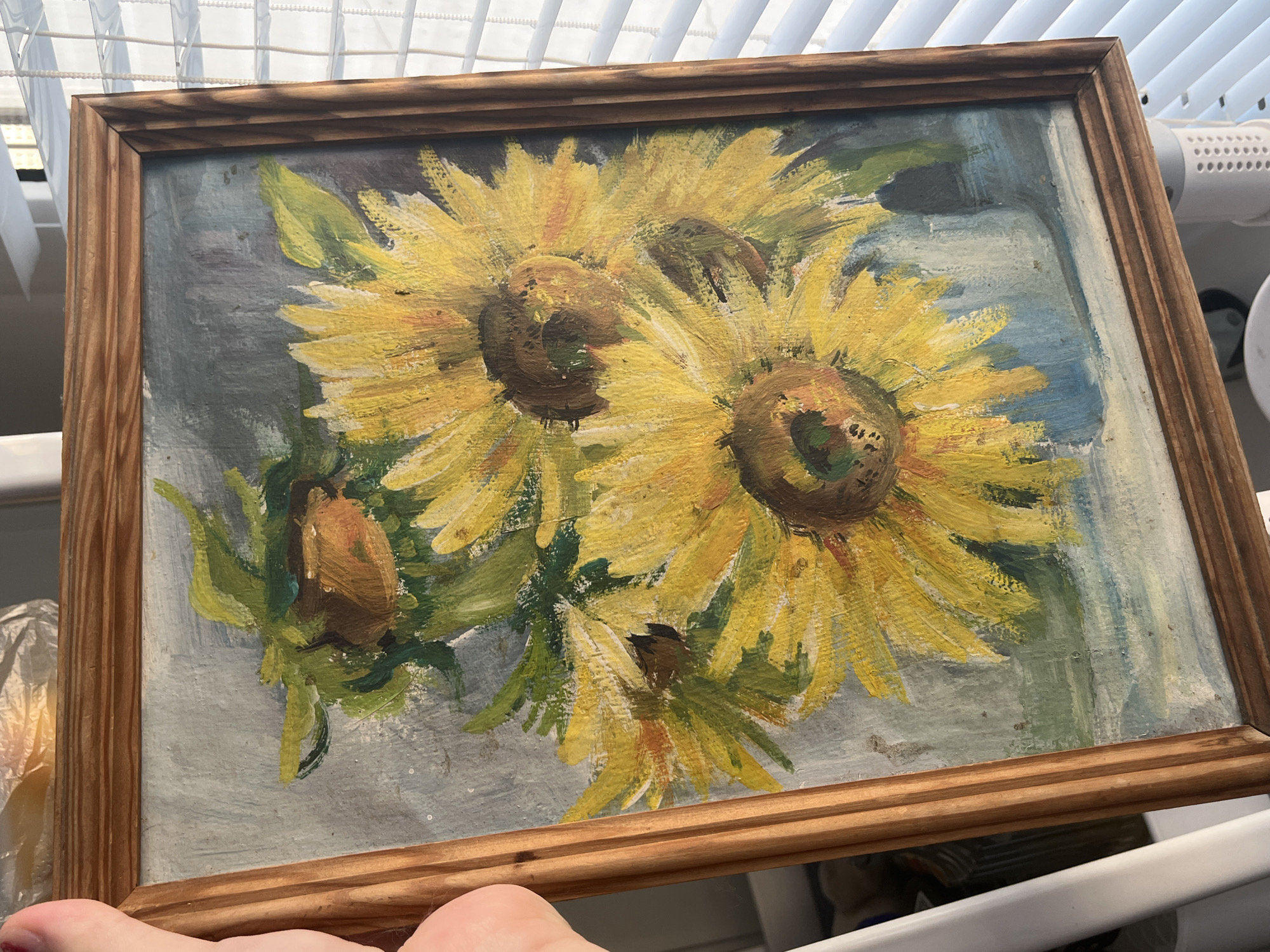 A framed oil painting of sunflowers is displayed. The painting depicts three prominent sunflowers in various stages of bloom, with vibrant yellow petals and brown centers. Green leaves and stems are also visible, arranged in a vase that appears to be light blue. The background is a muted grey with hints of blue and white, creating a soft, slightly textured effect. The wooden frame is simple and light brown with a visible grain. The painting is held by a person's hand with visible fingernails, suggesting it is being presented or moved. The scene is illuminated by natural light, casting shadows and highlighting the texture of the paint. The environment beyond the painting is out of focus but suggests an indoor setting with blinds on a window.
