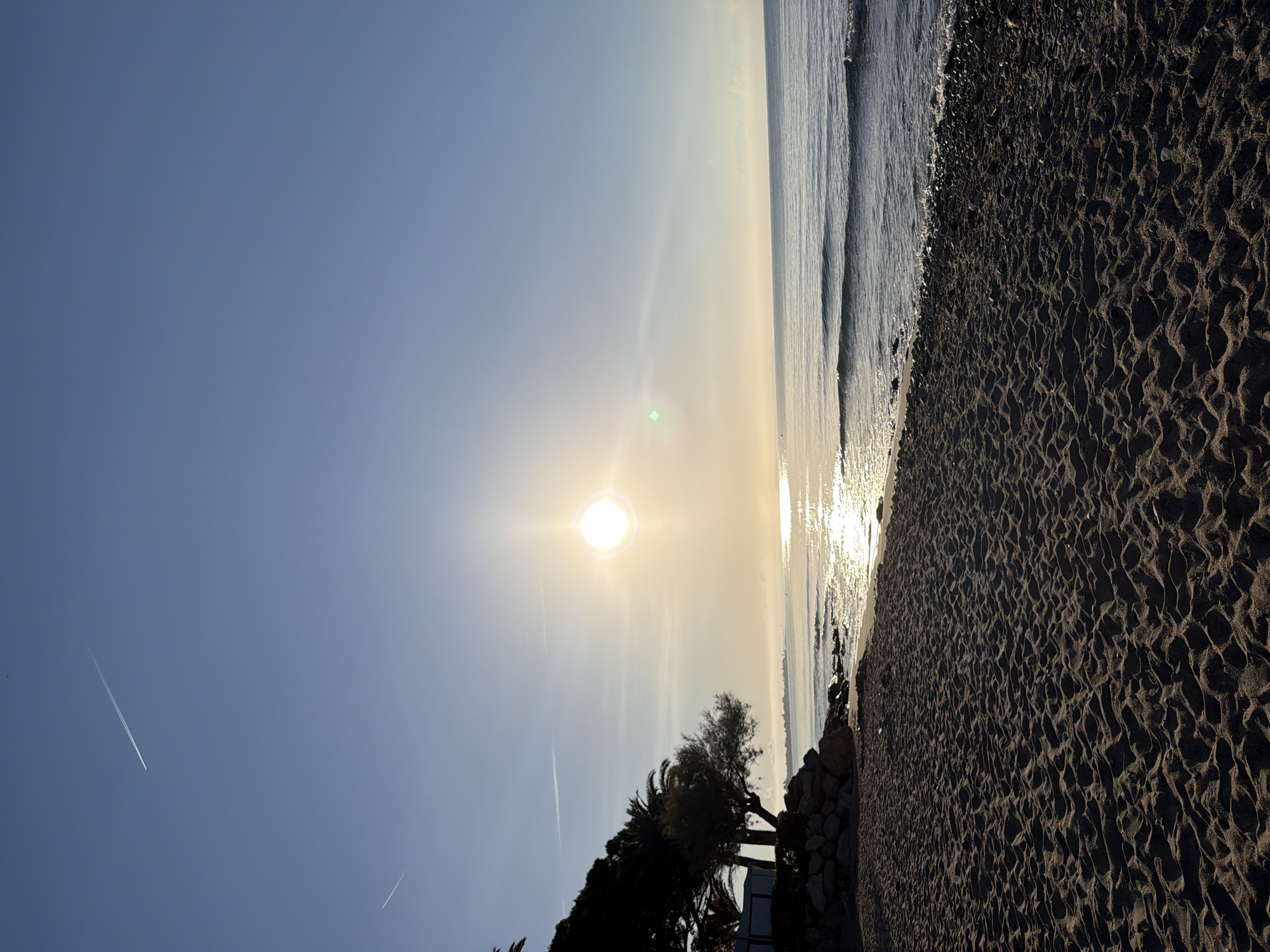 The image depicts a serene beach scene at Mont-roig del Camp, Spain, under a clear blue sky during sunrise. The sun, a bright orb, is positioned slightly left of the center, casting a warm glow and creating a shimmering reflection on the gentle waves of the Mediterranean Sea. The foreground is dominated by textured sand, showing the patterns left by receding tides, and a rocky shoreline to the left. A line of trees, including palm trees, marks the transition from the beach to the land. In the sky, two distinct contrails from airplanes are visible. The overall atmosphere is tranquil and bathed in the soft light of early morning.
