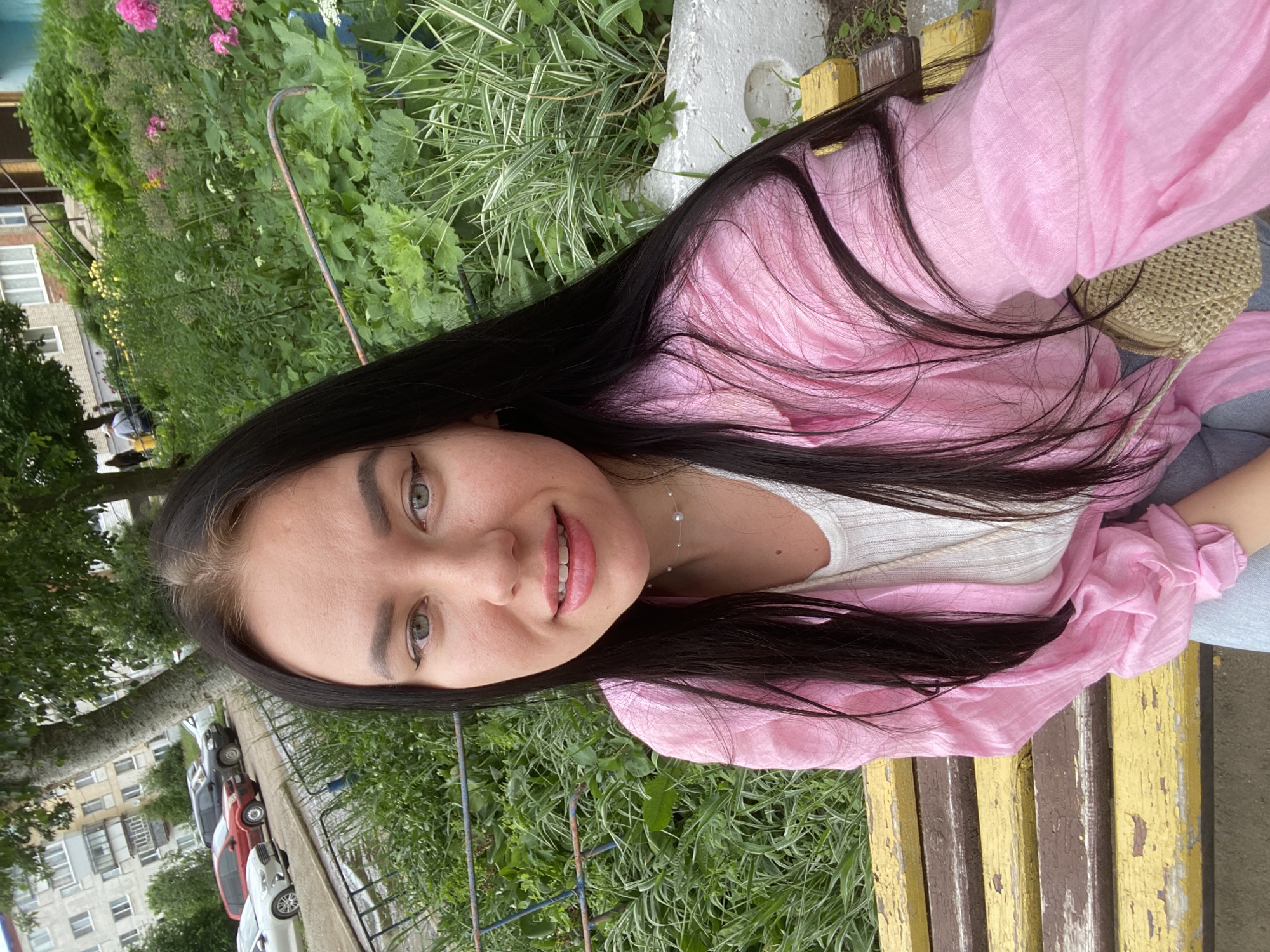 This is a portrait-oriented, slightly tilted, close-up photograph of a young woman. She has long, dark hair and is wearing a pink top over a white garment, and blue jeans. She is smiling and looking at the camera. She is sitting on a yellow and brown wooden bench outdoors. The background consists of lush green foliage and some flowering plants, with a hint of a building and parked cars visible on the left. The lighting suggests it is daytime. The overall mood is relaxed and casual. There is no visible text in the image.
