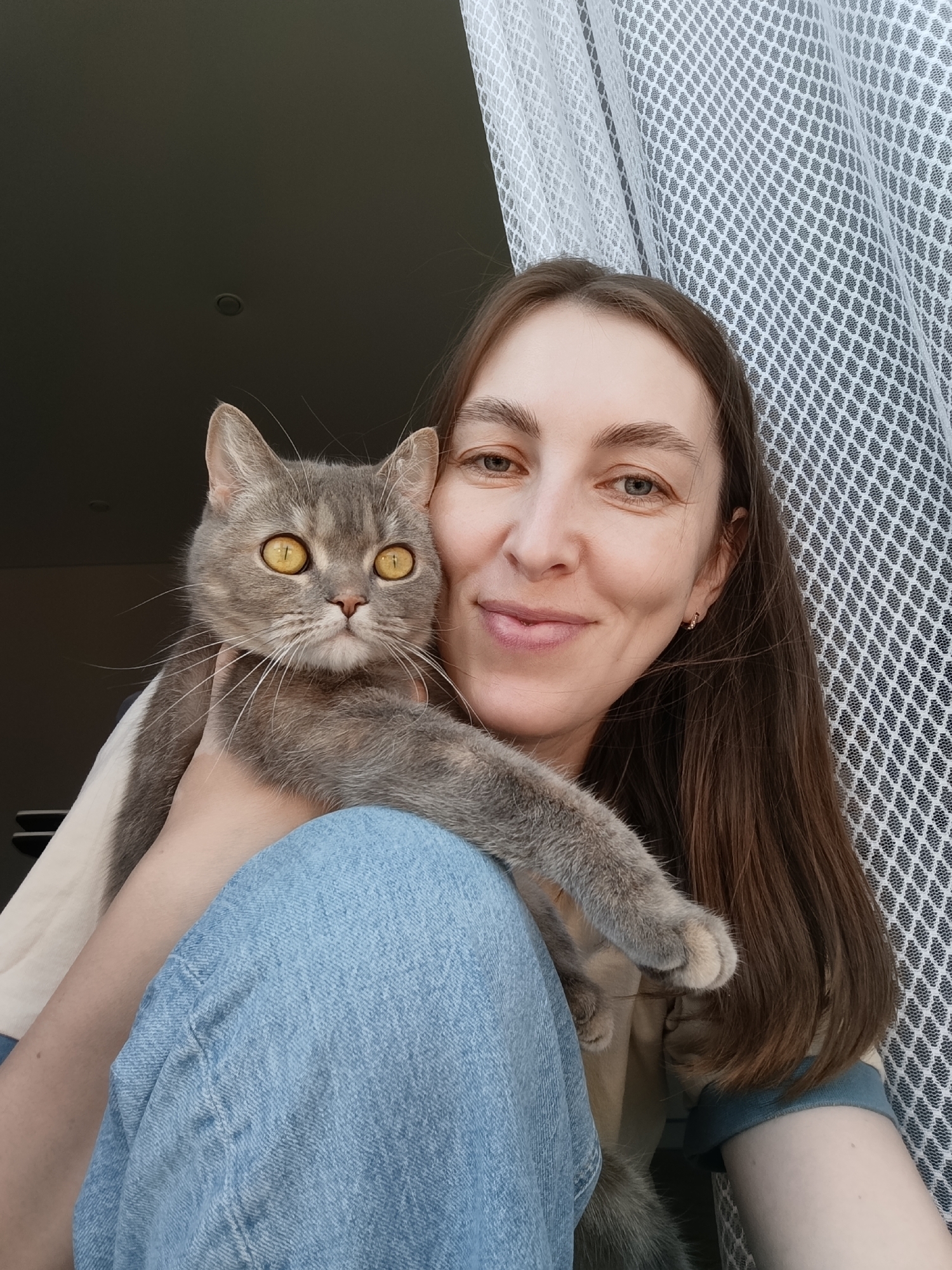 A woman with long brown hair, wearing blue jeans and a light-colored top, holds a gray tabby cat with yellow eyes. The background includes a partially visible window or doorway, and a white, textured curtain. It is not possible to determine the exact location of this image without additional information. It cannot be verified if this is in the city of Chaikovsky, Russia.