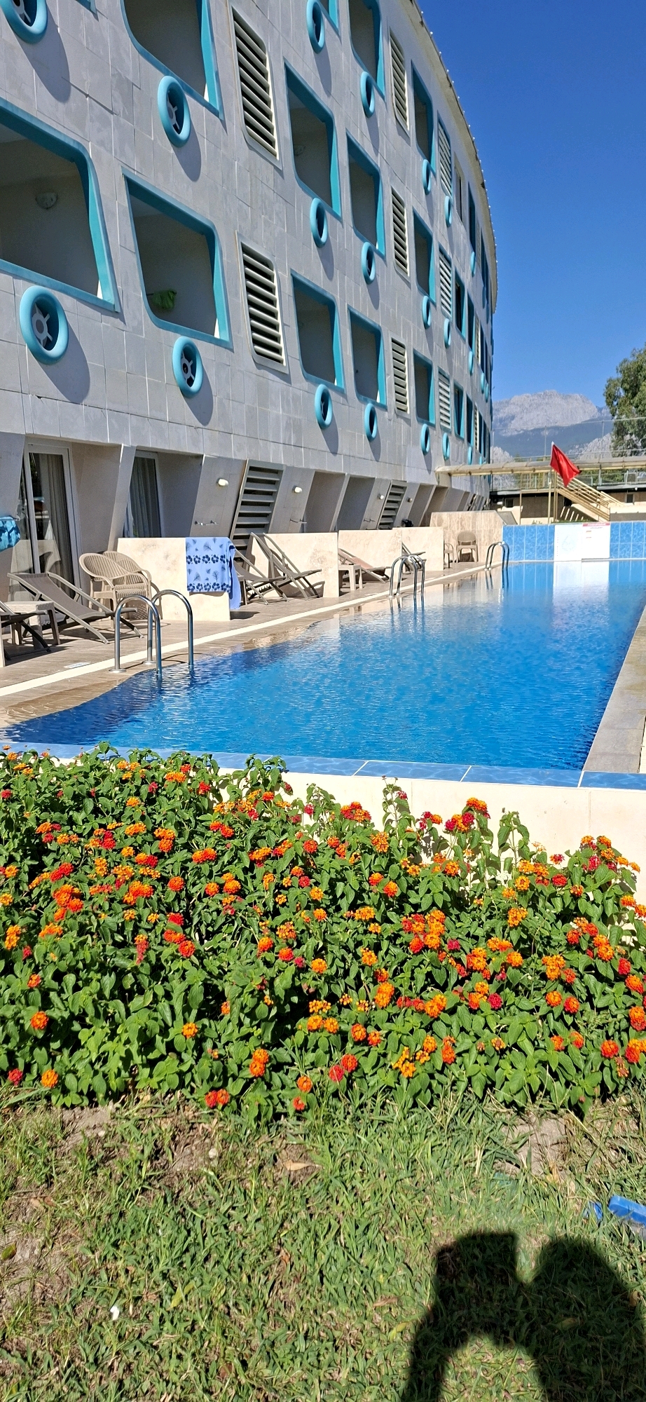 The image shows a swimming pool area at a resort in Kemer, Turkey. A modern building with an unusual architectural design, featuring numerous square windows with blue frames and circular blue vents, dominates the background. The building appears to be tiled in a greyish material. In front of the building, a bright blue swimming pool with metal ladders is visible. A tiled walkway lines the pool, with several lounge chairs and a small table set up. In the foreground, a vibrant hedge of orange and red flowers blooms. The ground in front of the flowers is covered with green grass, and the shadow of two people is cast on it. In the distance, mountains can be seen under a clear blue sky. A red flag is also visible on a pole near some stairs in the background.