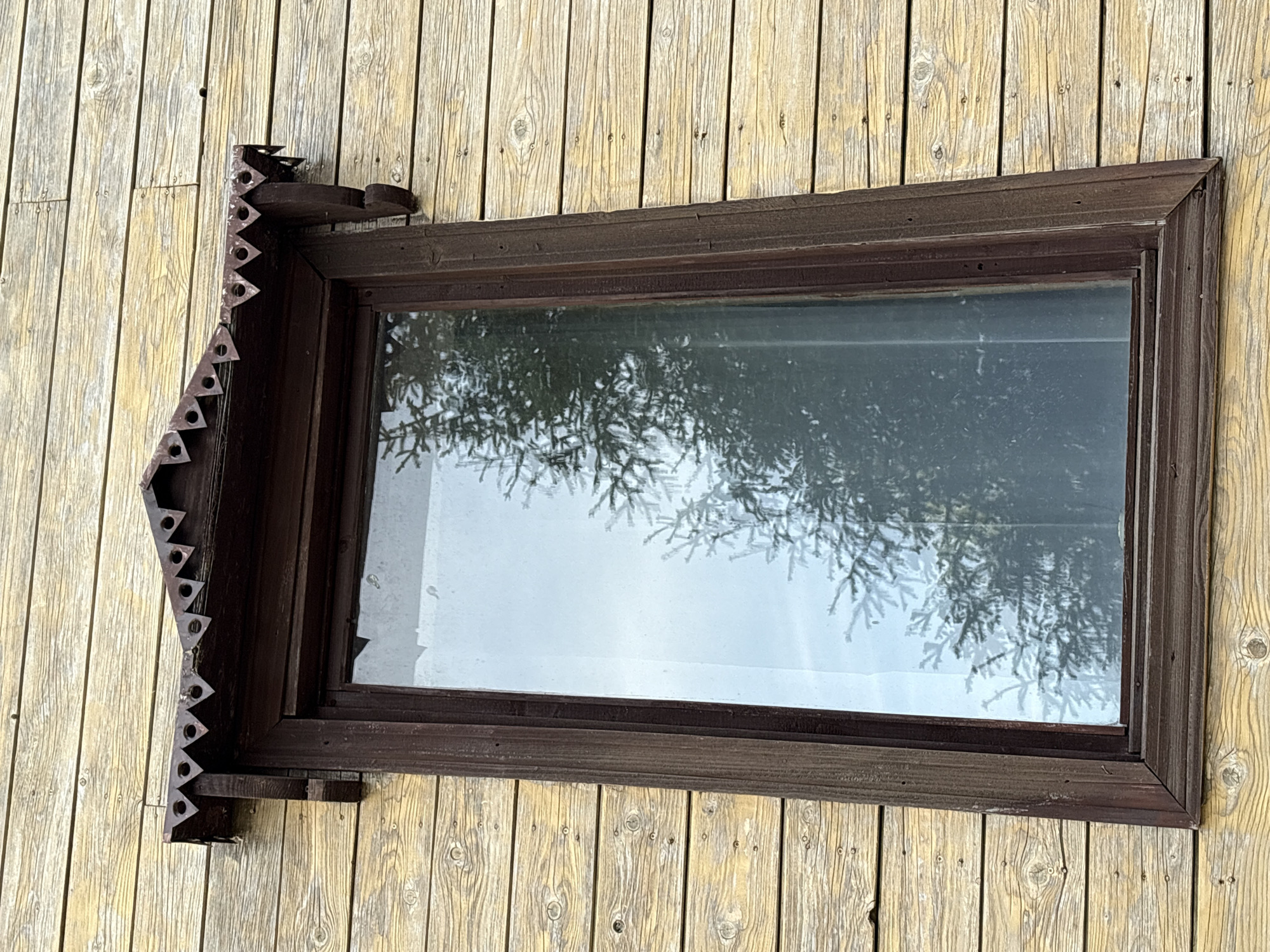 A horizontal, rectangular window with a dark brown frame is mounted on a weathered, yellow wooden wall. The window has decorative trim with triangular cutouts along its left side. Reflections of trees are visible in the window glass. The location is Zavod Mikhailovskii, Russia.