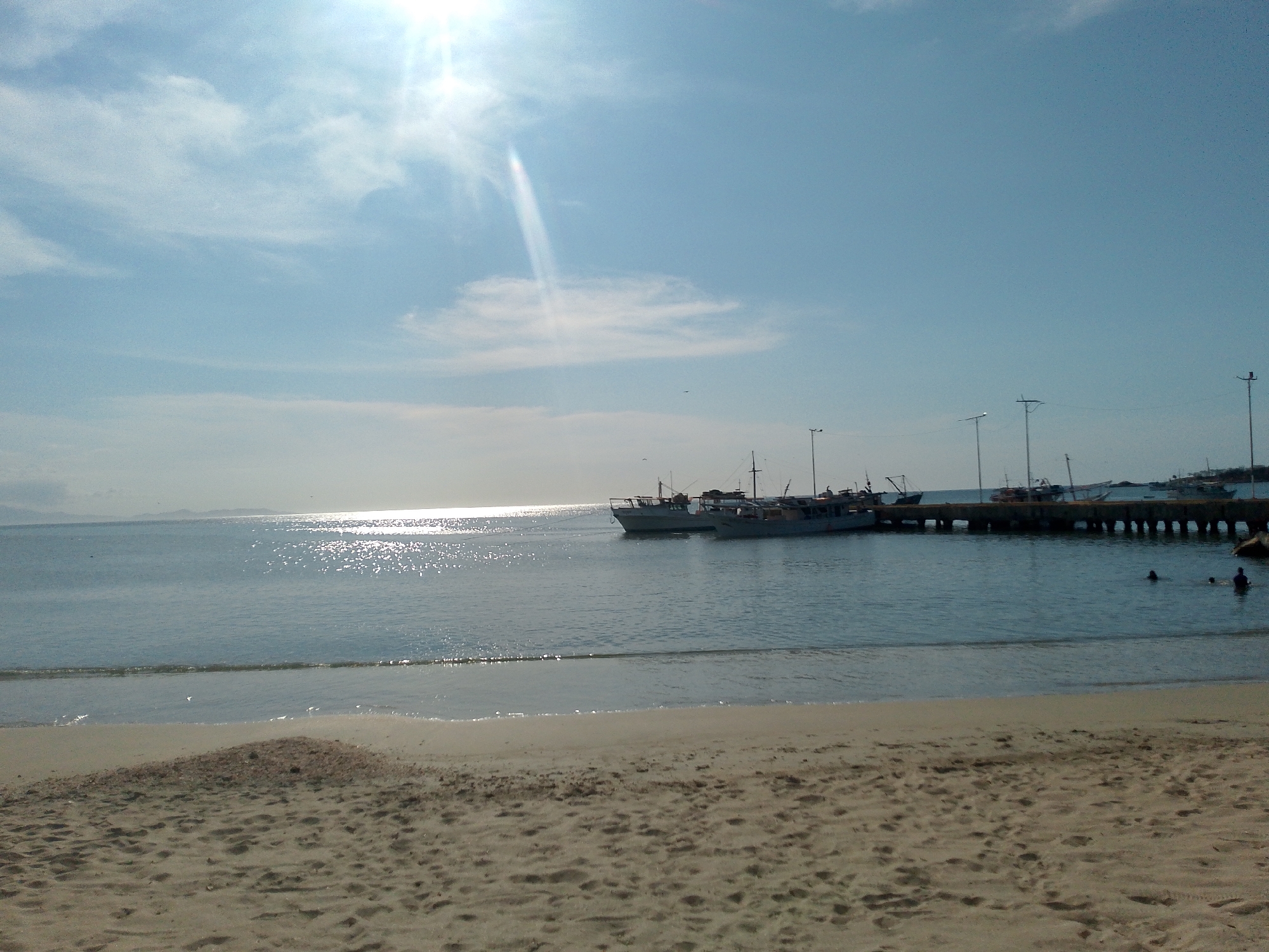 The image shows a beach scene in Juan Griego, Venezuela. The foreground is a sandy beach with visible footprints and patterns in the sand. The water is calm with sunlight reflecting on the surface. In the midground, there is a pier with several boats docked alongside. A few people are visible in the water, presumably swimming. Utility poles are erected along the pier. The background shows a light blue sky with some clouds, and the sun is shining brightly, creating a lens flare effect. In the far distance, a landmass is faintly visible.
