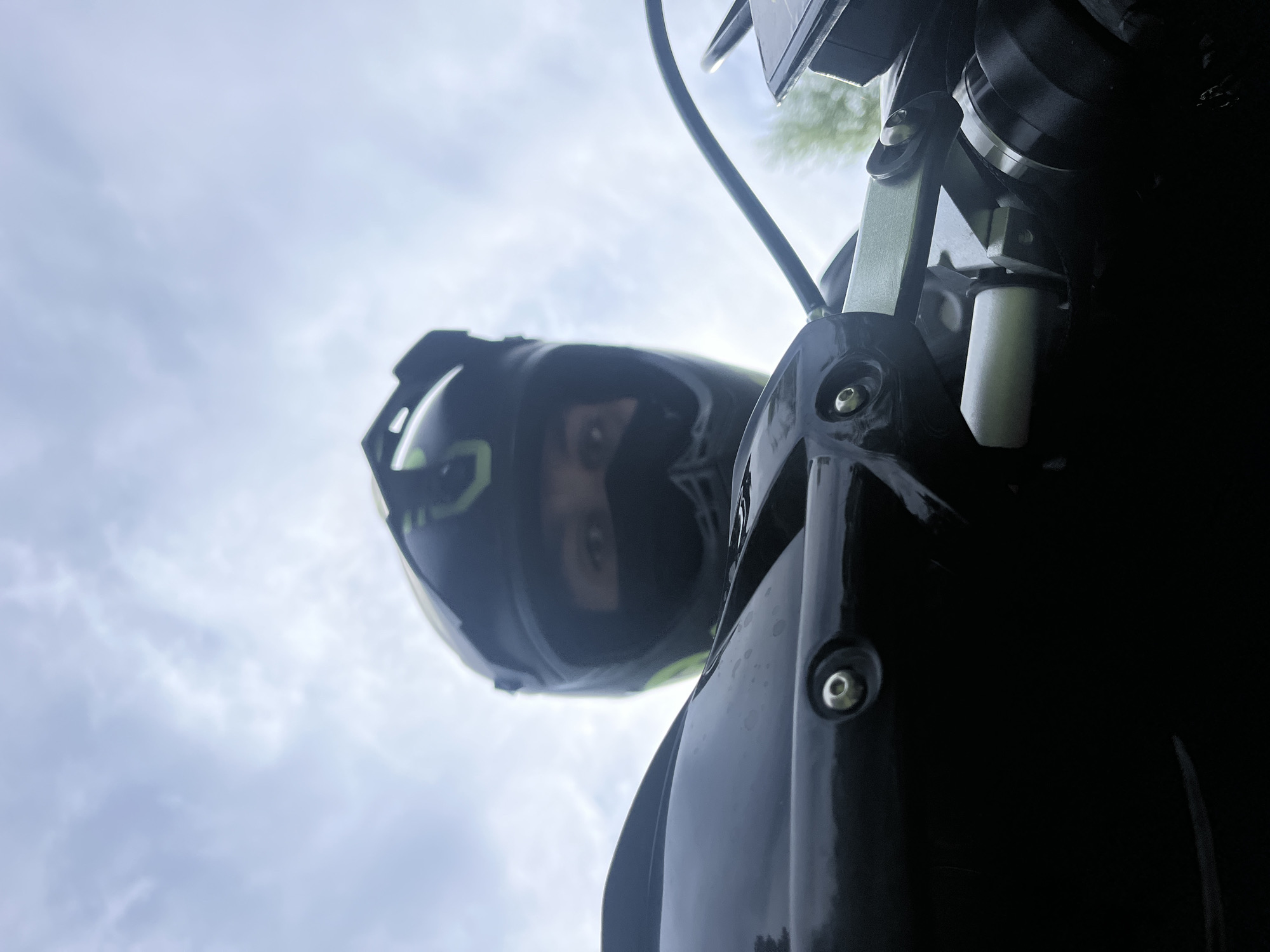 The image shows a person wearing a motorcycle helmet, looking down towards the camera. The helmet is black with green accents. The person's eyes are visible through the helmet's visor. The background is a cloudy sky. Part of a motorcycle is in the foreground, including a headlight and suspension components. The location is unspecified and cannot be determined from the image.