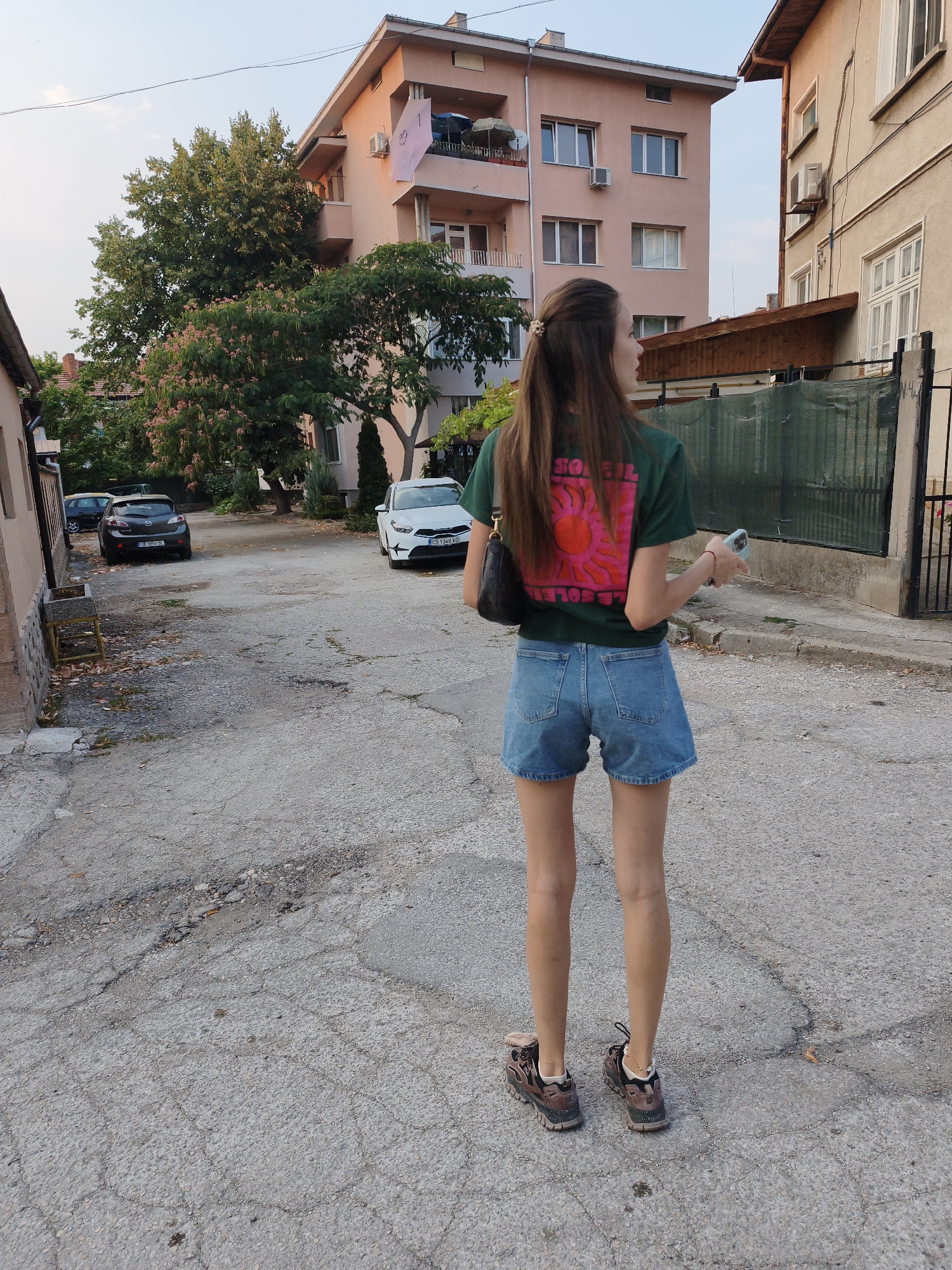 A young woman stands on a cracked asphalt street in Lovech, Bulgaria, facing away from the camera and looking to her right. She has long, dark hair pulled back with a gold clip. She is wearing a green t-shirt with a pink graphic of a sun and the text "SOLEIL" on the back, and denim shorts. She carries a black purse over her left shoulder and holds a light blue phone in her right hand. Her legs are bare, and she is wearing brown and black athletic shoes with a thin gold anklet on her left ankle. In the background, there are residential buildings, trees with pink flowers, parked cars including a white sedan and a dark gray hatchback, and a green fence. The sky is a pale blue, suggesting it is either early morning or late afternoon.