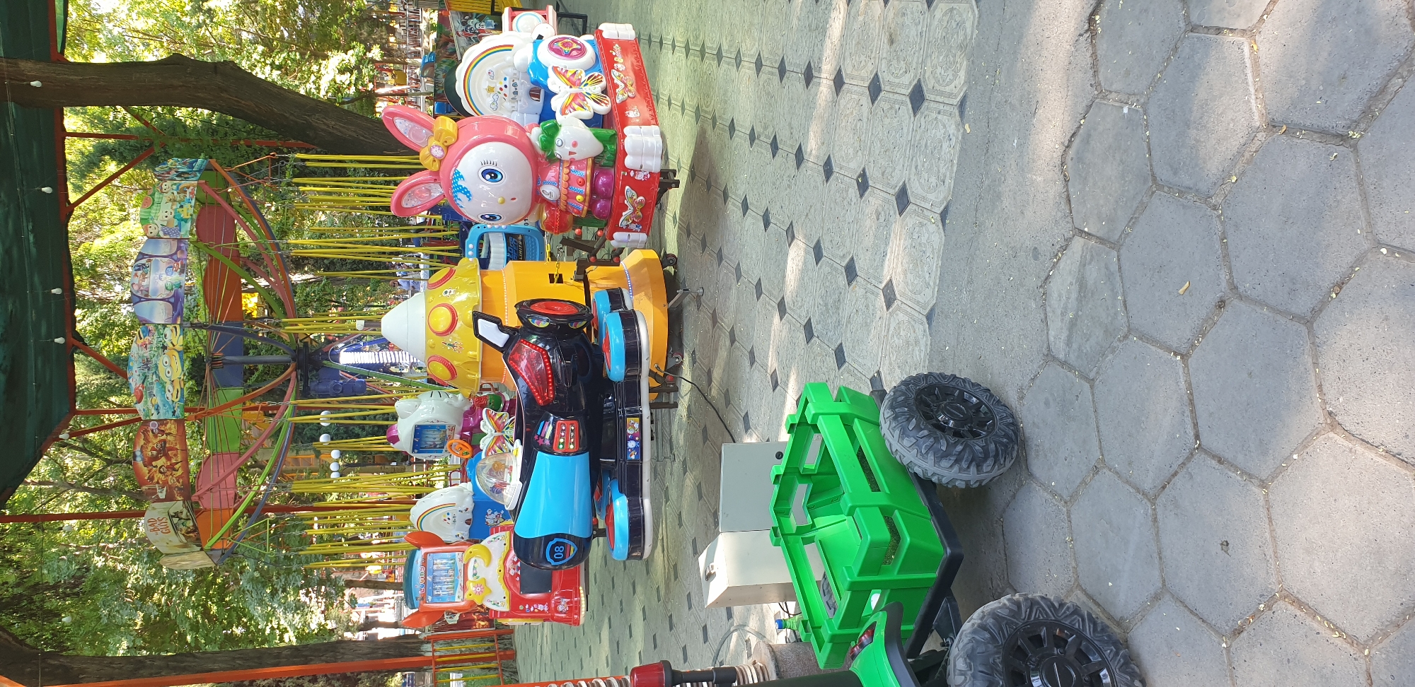 The image captures a section of a brightly colored amusement park ride, likely designed for children, situated outdoors on a paved ground. In the foreground, a green plastic wagon with large, rugged black tires is parked. Behind the wagon, several child-sized amusement rides are visible. These rides feature cartoon characters, a flying saucer-like structure, and a pink rabbit character. The rides appear to be attached to a central carousel-like structure with yellow poles and a colorful canopy overhead. The background reveals a dense area of trees, suggesting a park or green space within the city of Fergana, Uzbekistan. The lighting indicates it is daytime, with sunlight filtering through the trees and casting shadows on the ground. There are no people clearly depicted in the image.