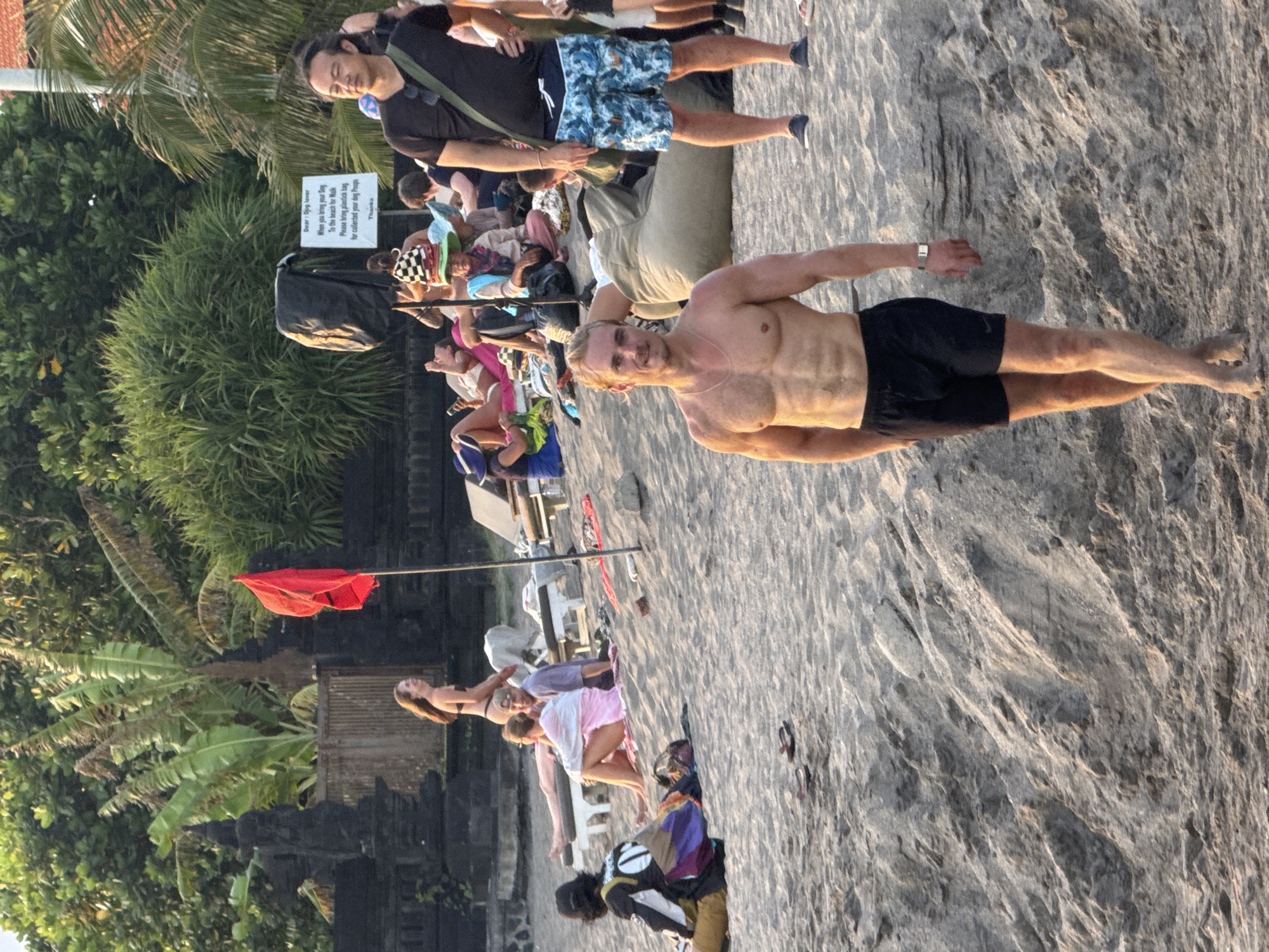 This image captures a sunny day on a beach, possibly in the city of North Kuta, Indonesia, based on the tropical foliage and architectural elements. A fair-skinned man with a muscular build is standing bare-chested in black shorts on the sand, looking directly at the camera.
Behind him, several individuals are relaxing on the beach, including groups lounging on chairs and towels. A red flag on a pole is visible, possibly indicating a caution area or a lifeguard station. Further back, a person wearing shorts is carrying a bag.
To the left, a building with traditional architecture and lush tropical trees is present. A sign for dog owners is placed on the opposite side. The sand appears coarse and light-colored.