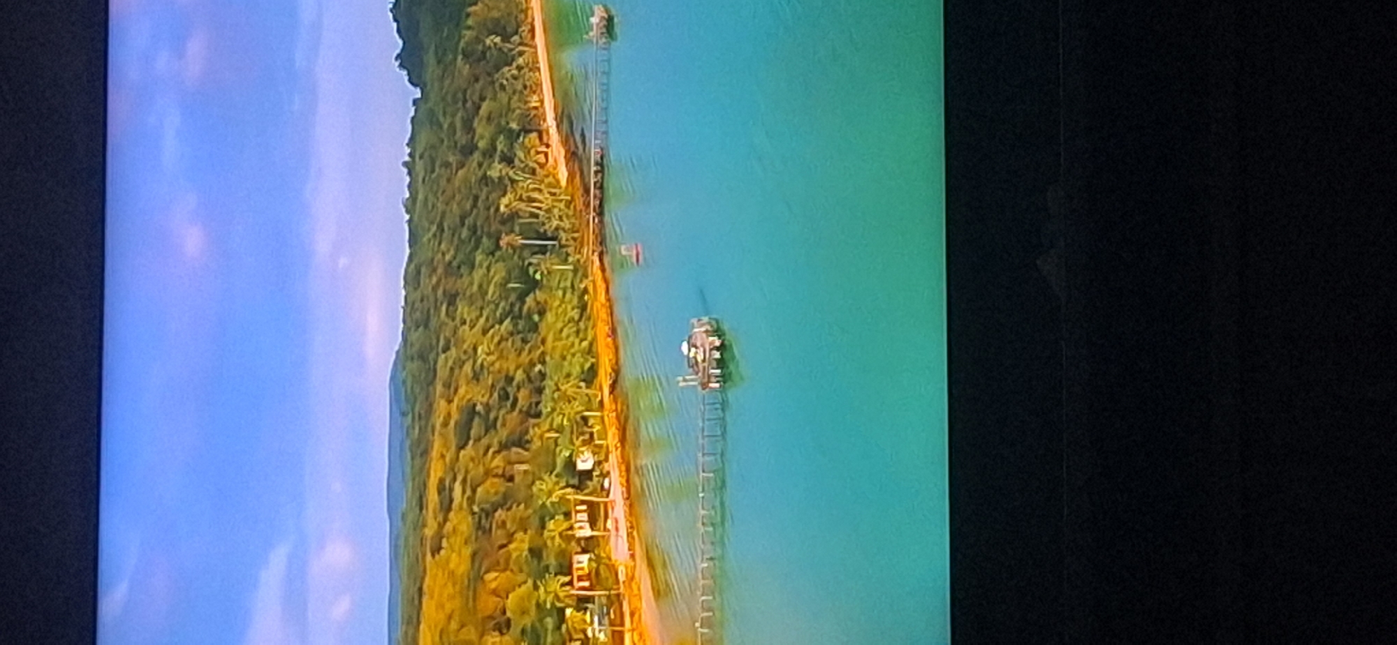 The image shows a tropical coastal scene with a clear blue sky and turquoise water. A lush, green, tree-covered hillside slopes down to a sandy beach lined with palm trees and small buildings. A wooden pier extends from the shore into the water, leading to a small circular structure. Another smaller pier is visible further down the coast. There are no people or visible activity in the scene. The location is not Bila Tserkva, Ukraine, as the scenery is clearly tropical and coastal.