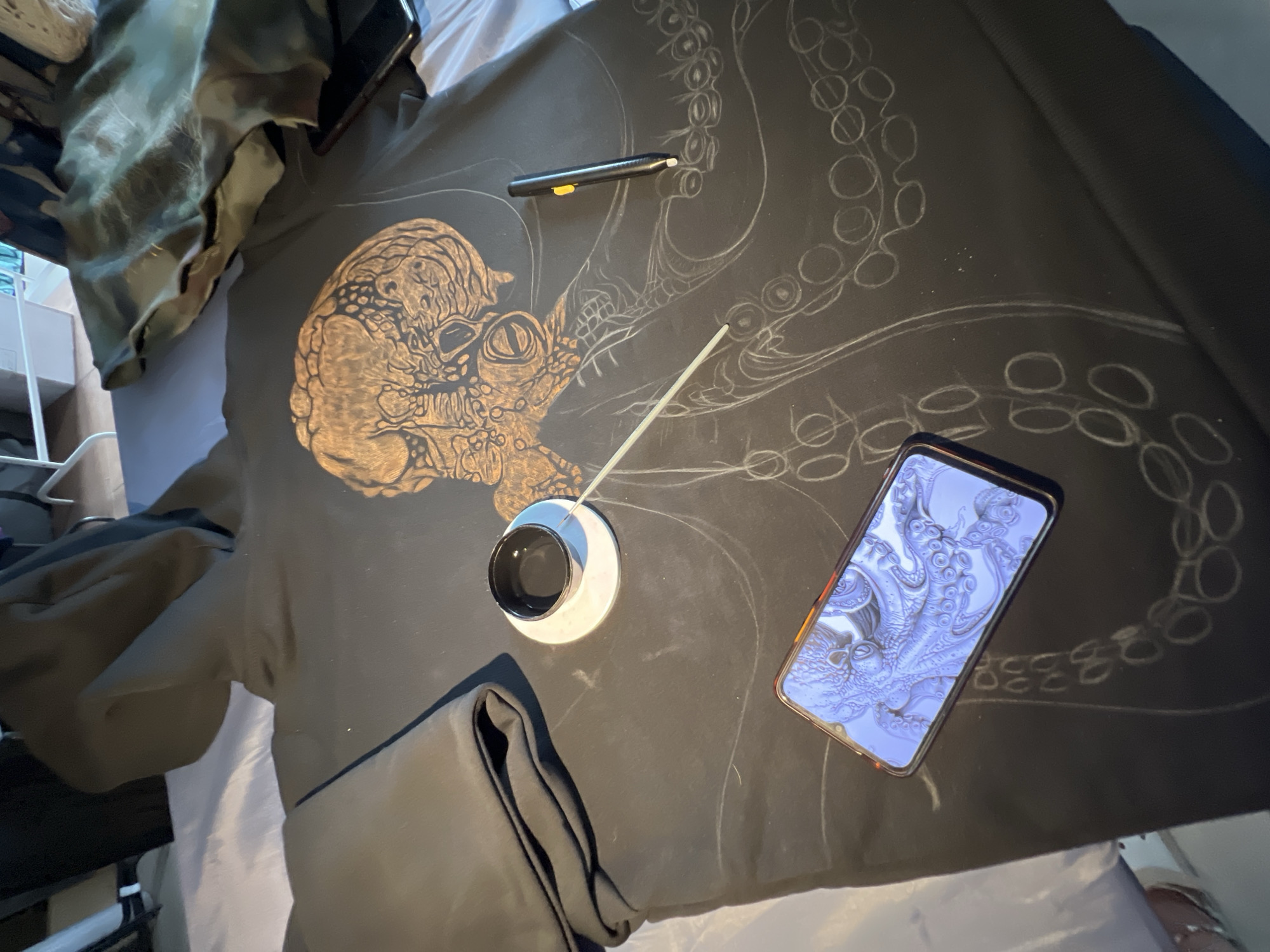 The image shows a black hooded sweatshirt laid out on a bed. The sweatshirt has a large illustration of an octopus being drawn on it with white chalk or a similar marking tool. The head of the octopus is already drawn and filled in with a golden-brown color, while the tentacles are still in the sketching phase. A mobile phone is placed on the sweatshirt, displaying an image of an octopus, likely for reference. Next to the phone is a small white saucer with a black cup or inkwell on it, and a long white stick, possibly a drawing tool. A black stylus pen with a yellow button is also visible on the sweatshirt. In the background, a folded dark green garment is placed to the left of the sweatshirt, and other items are visible on a bed in a dimly lit room. The image does not contain any specific landmarks or details that would allow for the identification of its location in Petrozavodsk, Russia.