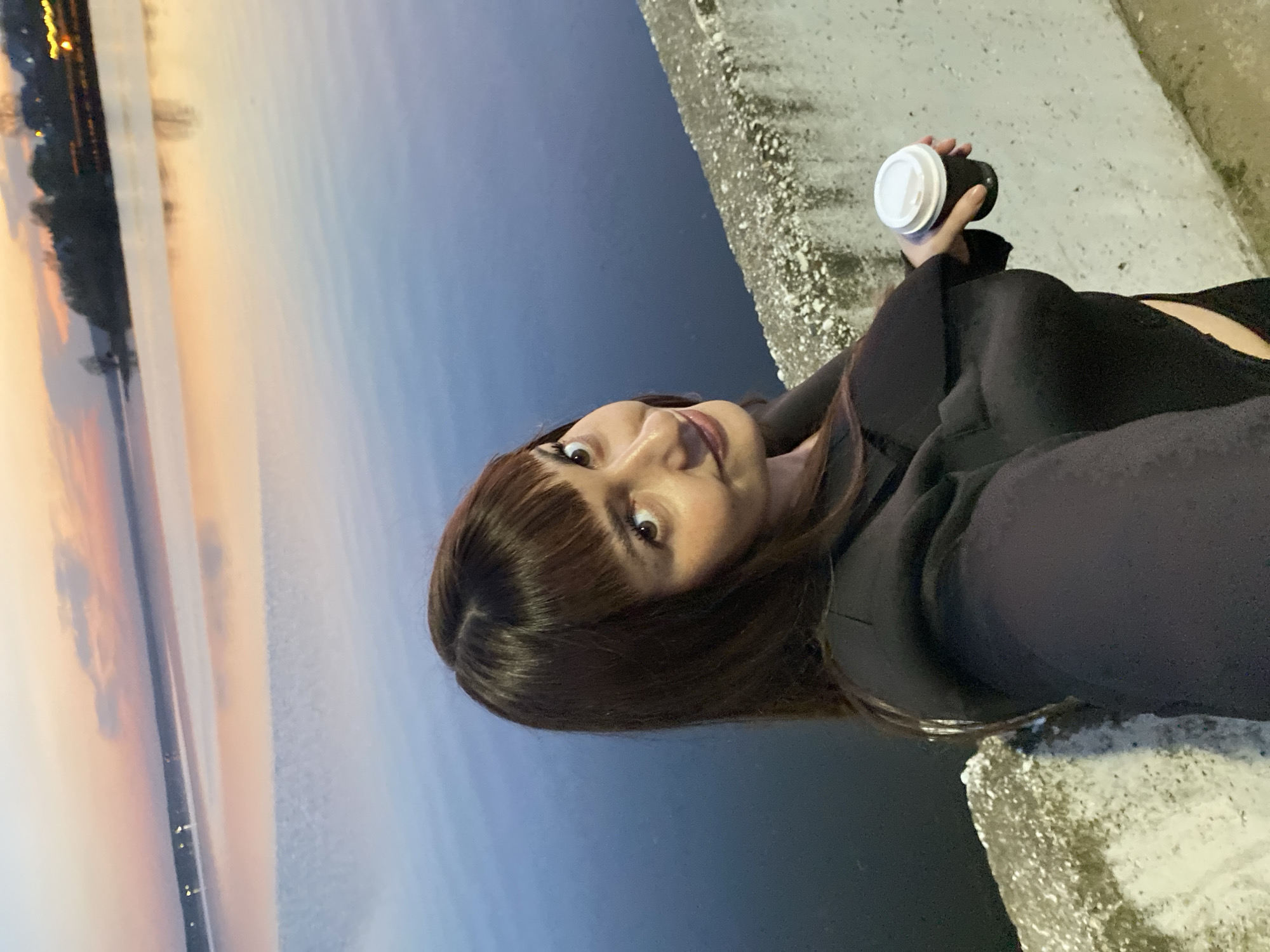 A young woman takes a selfie while holding a disposable coffee cup with a white lid. She has brown hair with bangs and is wearing a black top or jacket. Behind her is a body of water reflecting a soft, pastel-colored sunset, possibly indicating early morning or late evening. The scene is situated near a concrete embankment or wall, with a slightly rough, textured surface. The location appears to be outdoors and likely near the Votkinsk Reservoir, based on the sunset reflecting on water.