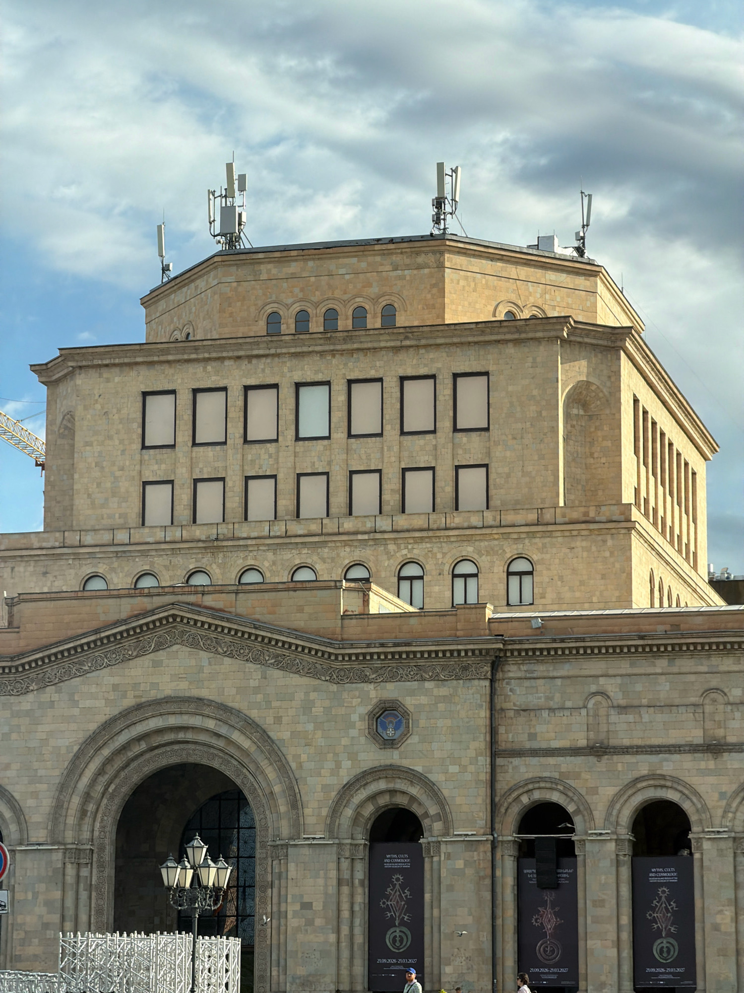 The image shows a close-up, exterior view of the National Gallery of Armenia, located in Republic Square, Yerevan, Armenia. The building is constructed of light-colored stone, with a prominent multi-story facade featuring a series of arched entrances on the lower level and rows of rectangular windows on the upper floors. On the rooftop, there are several communication antennas and structures.
The sky above is partially cloudy, with a mix of blue sky and grey clouds. In the foreground, a vintage-style street lamp stands near the center of the image, and a white metal scaffolding structure is visible to the left. Several large banners are displayed under the arched entrances on the lower level of the building, featuring artistic designs and text. Two individuals are partially visible walking in front of the building on the right side of the frame. A construction crane is visible on the far left of the image, behind the building.