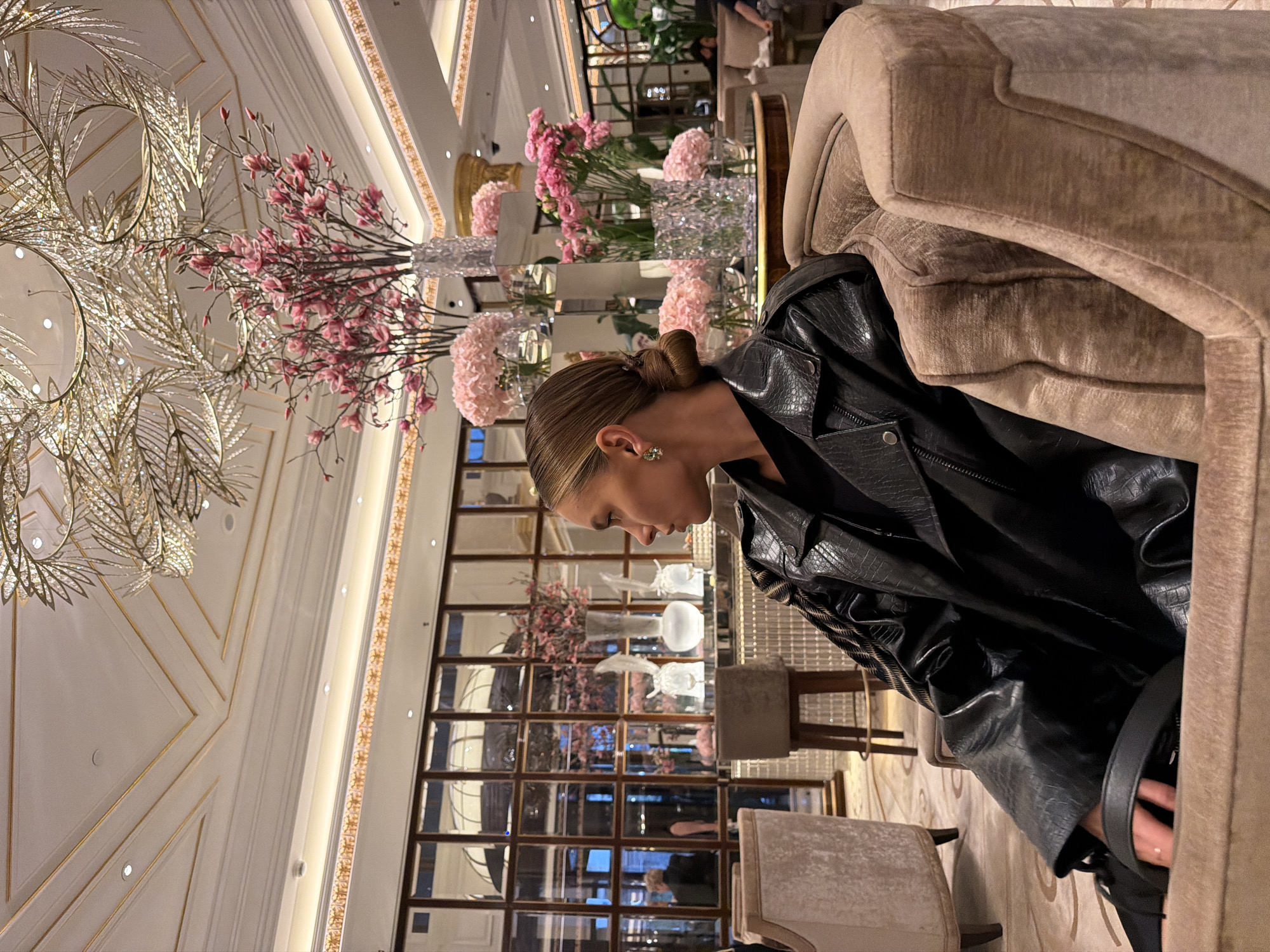 In this media file, a woman is seated in a plush, light-brown armchair in an opulent indoor setting. She is wearing a black leather jacket with a textured, crocodile-like pattern. Her hair is styled in a neat bun at the nape of her neck, and she has a delicate earring in her visible ear. The woman is looking down and to the side, appearing contemplative.
The background reveals a luxurious interior, characterized by a grand chandelier with crystal accents, ornate gold trim on the ceiling, and elegant floral arrangements of pink flowers in tall, crystal vases. Large mirrored panels or windows reflect the surroundings, creating a sense of depth and adding to the sophisticated ambiance. The lighting suggests it is evening or indoors with artificial light. The setting appears to be a high-end lounge, restaurant, or hotel lobby in Moscow, Russia, given the provided location context. The overall mood is one of quiet elegance and refinement.
