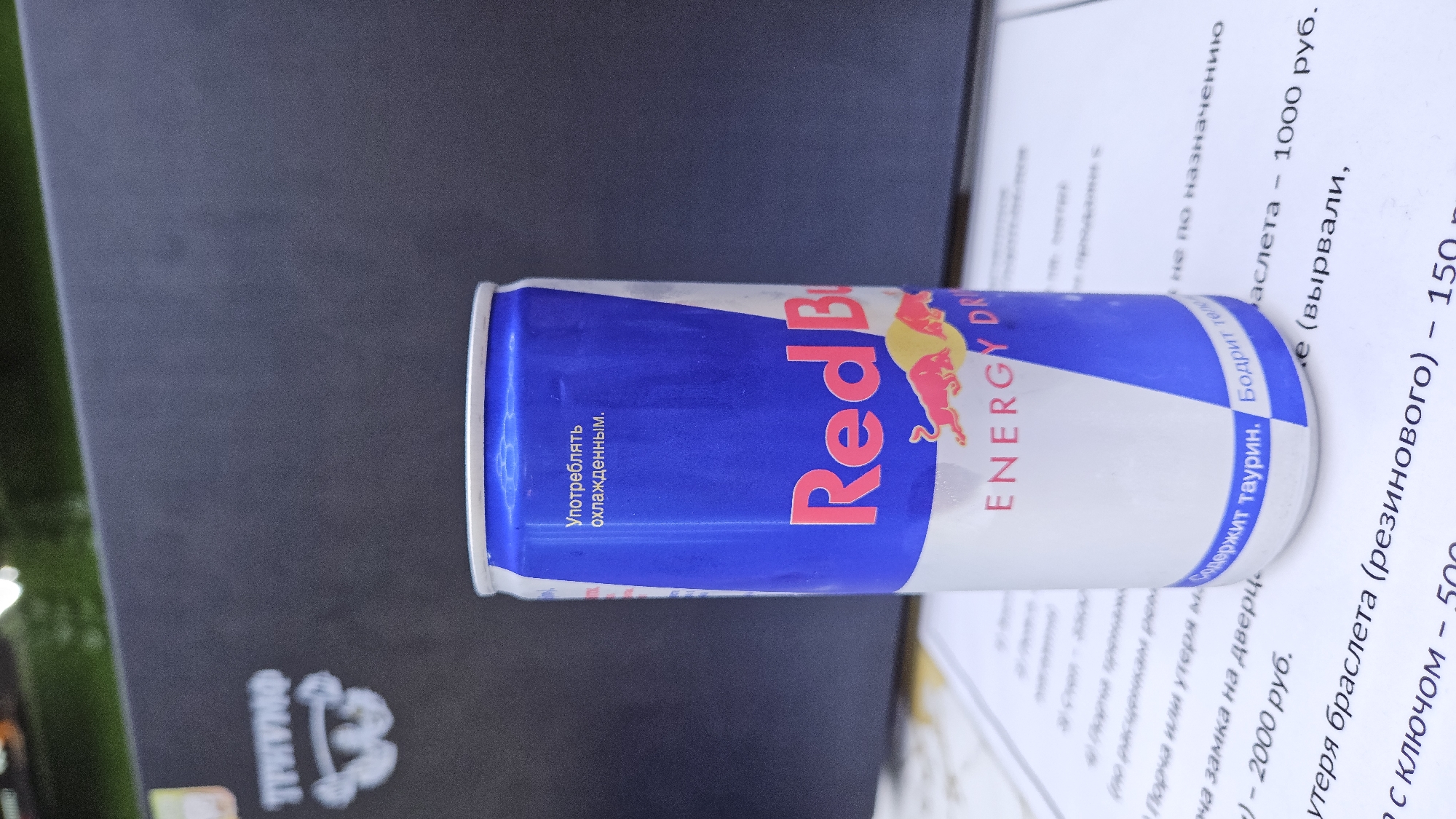A close-up shot features a blue and white Red Bull energy drink can resting on a piece of paper, partially obscuring some printed text written in Russian. To the left, the can is adjacent to a dark gray surface with a white logo. The Russian text on the can includes "Употреблять охлажденным." and "Red Bull ENERGY DR". Part of the can is resting on a white paper with Russian text. Some of the readable text includes "Содержит таурин. Бодрит тело", followed by what appears to be a price list in Russian rubles, mentioning items such as a bracelet for "1000 руб." and another item for "2000 руб." The image does not provide information about the specific location in Sevastopol, Ukraine.