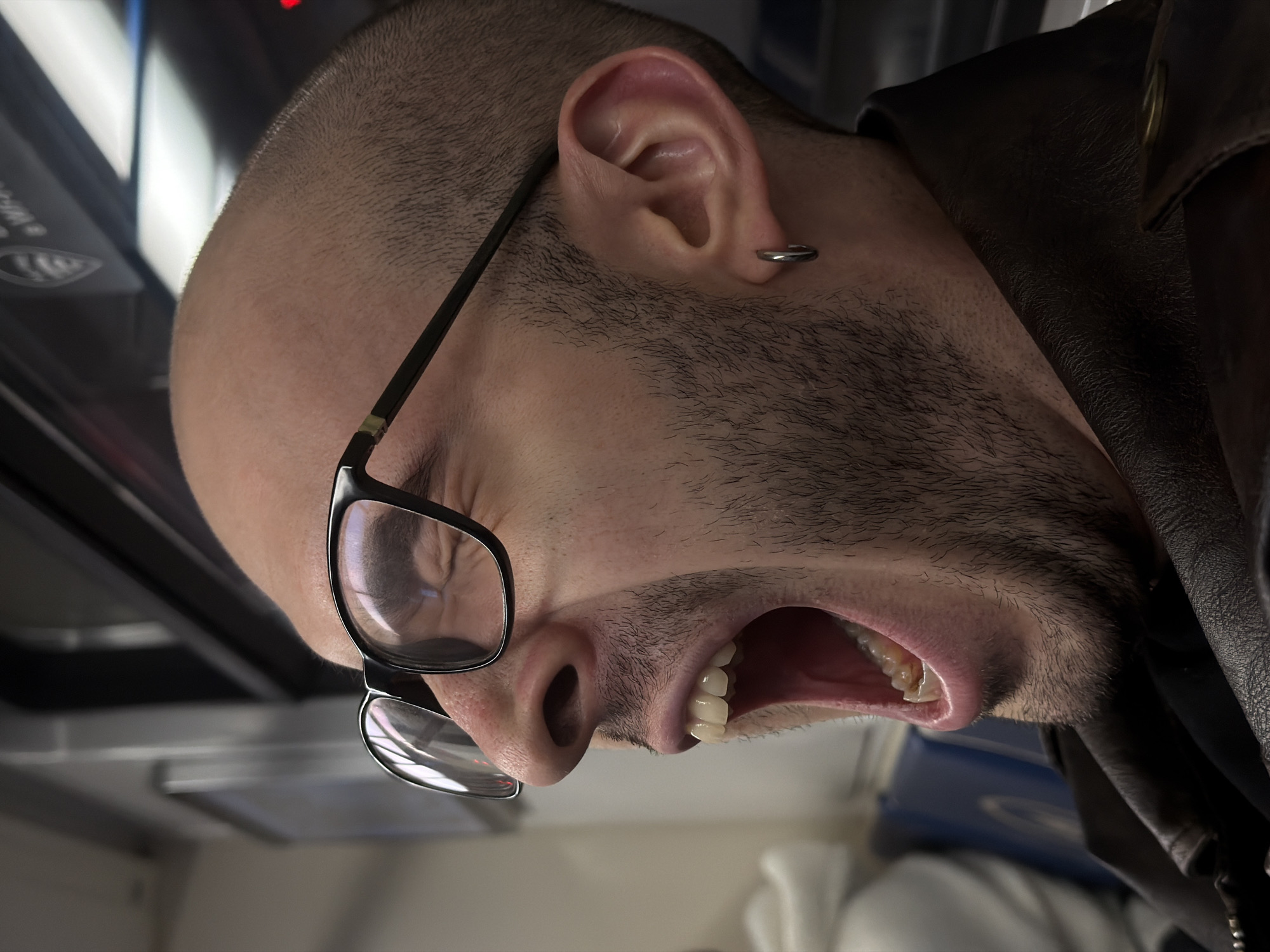 A close-up, low-angle shot depicts a man in what appears to be a vehicle, possibly public transport. He has a shaved head, a beard, and is wearing glasses. His mouth is open wide, seemingly in a yawn or shout. He also has a piercing in his ear. He is wearing a brown leather jacket. The background is blurry, showing the interior of the vehicle with elements like windows and seating. The exact location within Moscow cannot be determined from the image.
