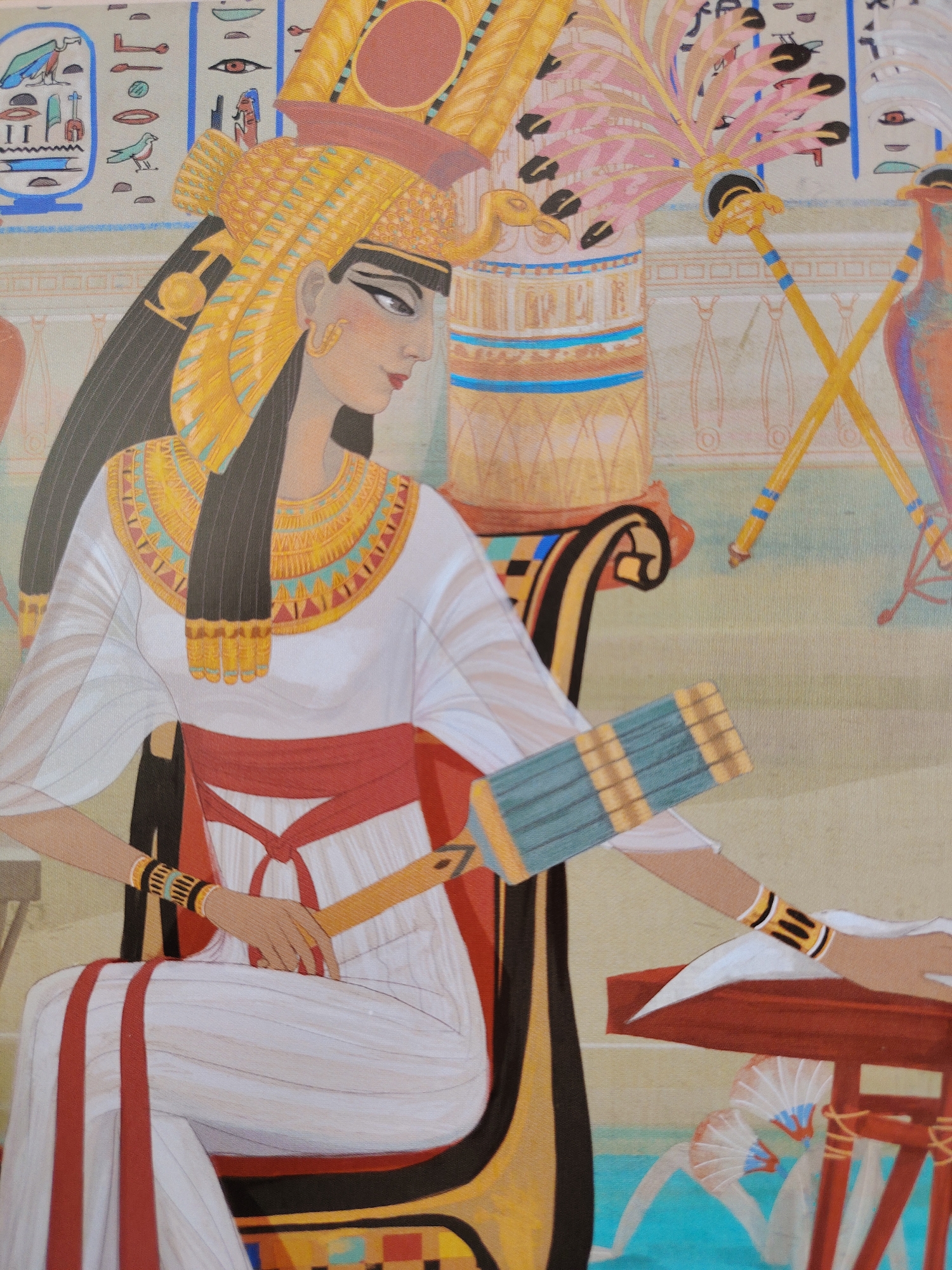 This image depicts a scene from ancient Egypt, rendered in a stylized, colorful illustration. The central subject is a woman, likely a queen or noble, adorned in elaborate Egyptian regalia. She wears a tall, golden headdress with ornate detailing and a large, circular amulet at the front. Her neck is encircled by a wide, golden collar inlaid with geometric patterns of turquoise, red, and blue. She is dressed in a flowing white robe with red sashes tied around her waist and draped over her shoulders. Her skin is fair, and her eyes are dramatically lined in black.
She is seated in a richly decorated chair with a dark, curved armrest and a base adorned with geometric patterns. Her left arm rests on the armrest, while her right arm is extended, holding a fan-like object with blue and gold slats. Another object, a golden staff, rests against her arm. The background suggests an interior setting within an ancient Egyptian palace or temple. Hieroglyphics are visible on the wall behind the figures, depicted in muted tones. To the right, two ornate golden staffs with feathered ornamentation are crossed, further emphasizing the regal atmosphere. Another figure, partially visible on the far right, appears to be a bird or a statue of a bird, rendered in vibrant orange and blue. The overall impression is one of opulence, ceremony, and historical depiction. The lighting is bright and even, suggesting daytime. There is no visible text.